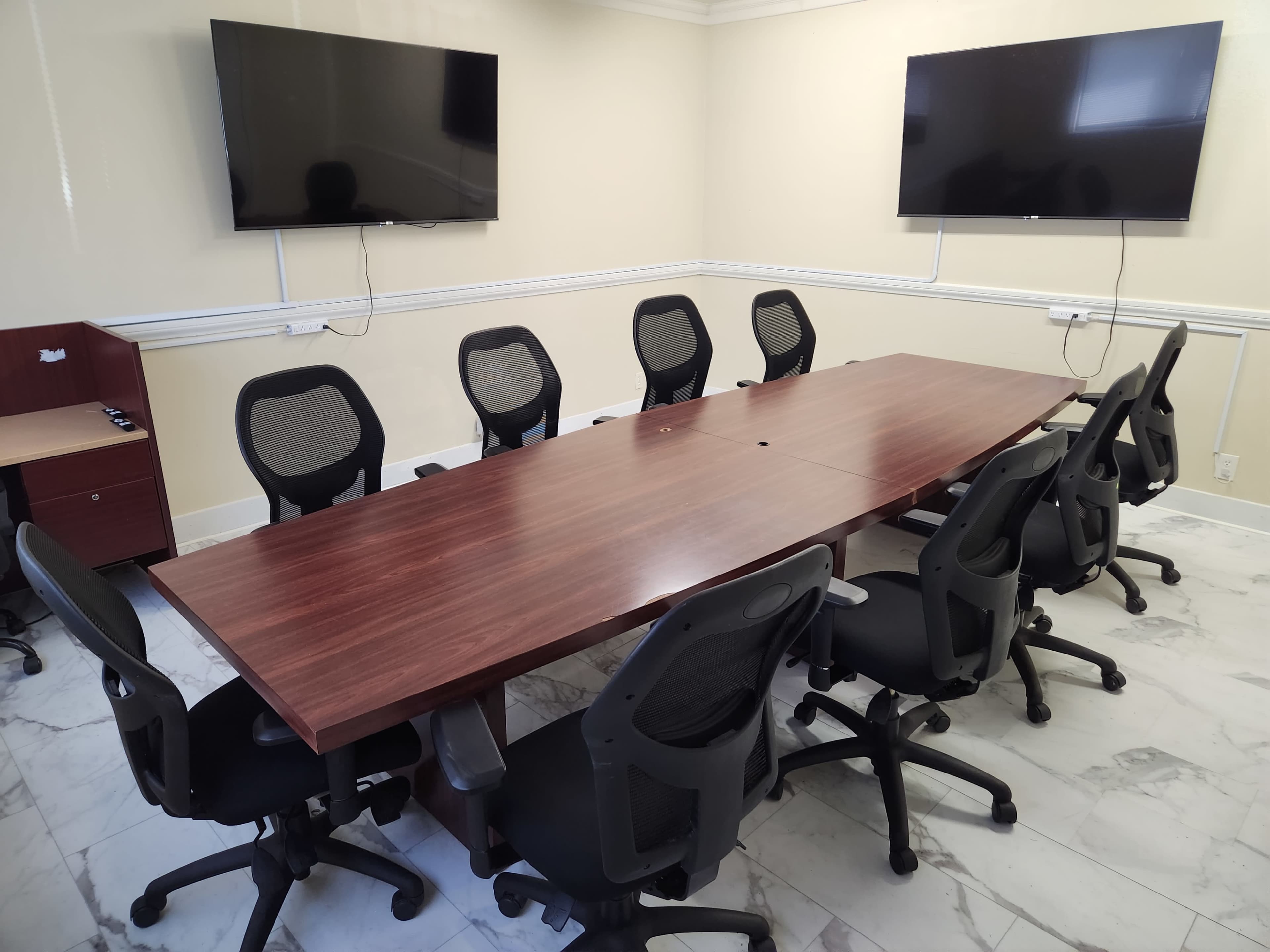A conference room is equipped with a large wooden table surrounded by ergonomic chairs and two mounted televisions on the walls.