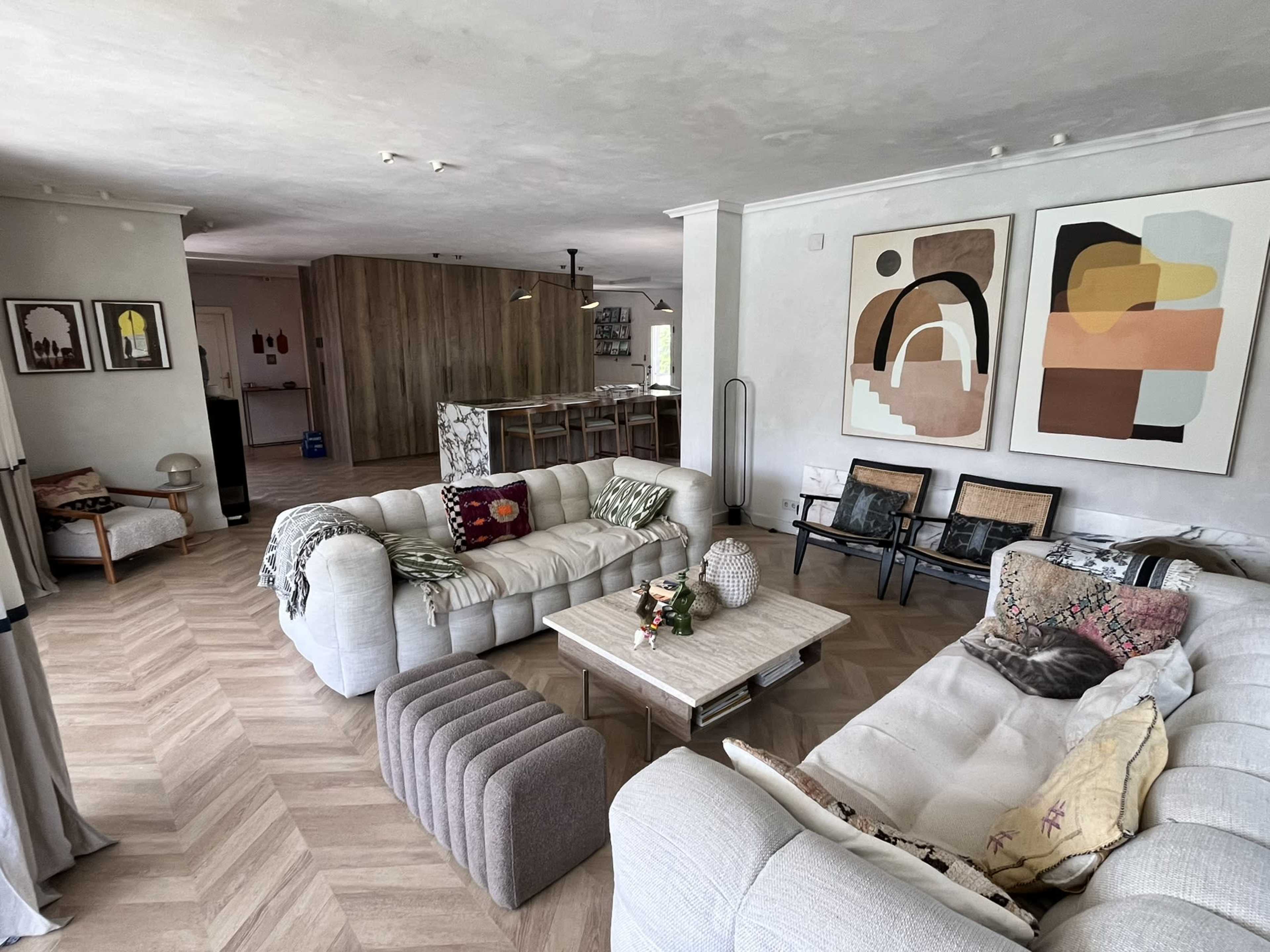 The image shows a spacious living room with light-colored furniture, abstract art on the walls, and a dining area in the background.