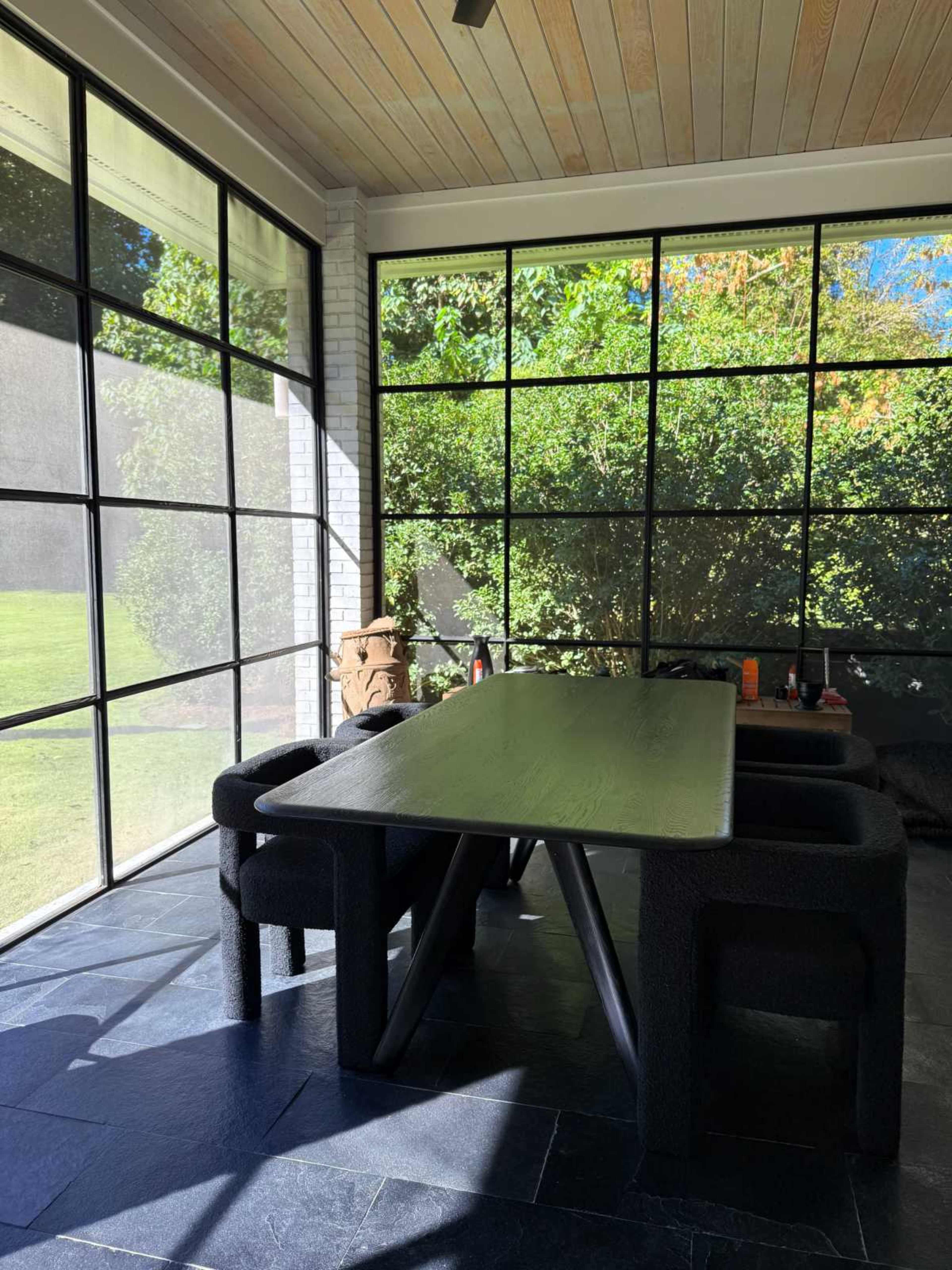 The image shows a spacious dining area with a large table and black upholstered chairs, surrounded by floor-to-ceiling windows that offer a view of green foliage outside.