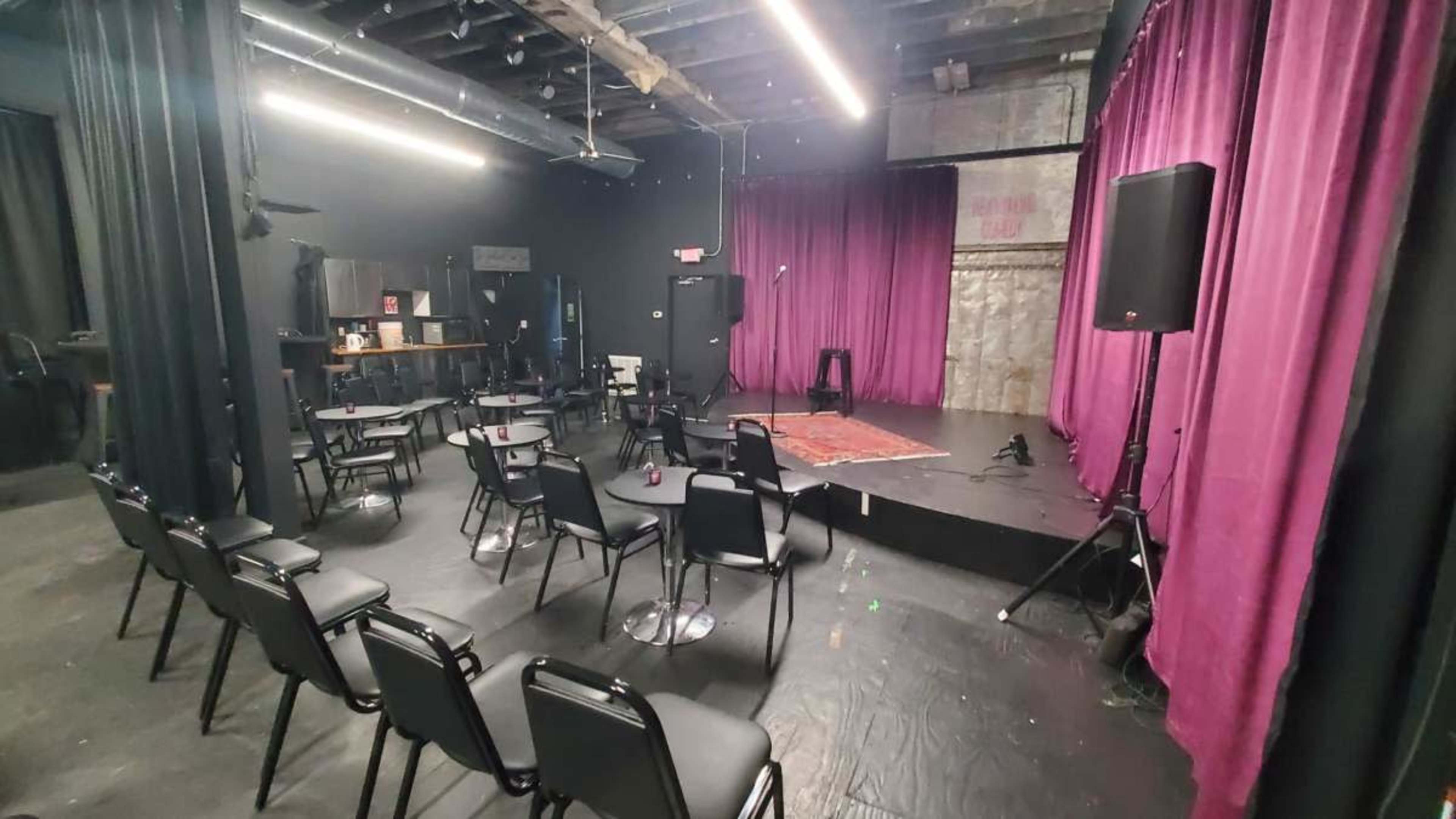 The image shows a small theater setting with empty black chairs arranged in front of a stage featuring purple curtains and a microphone.