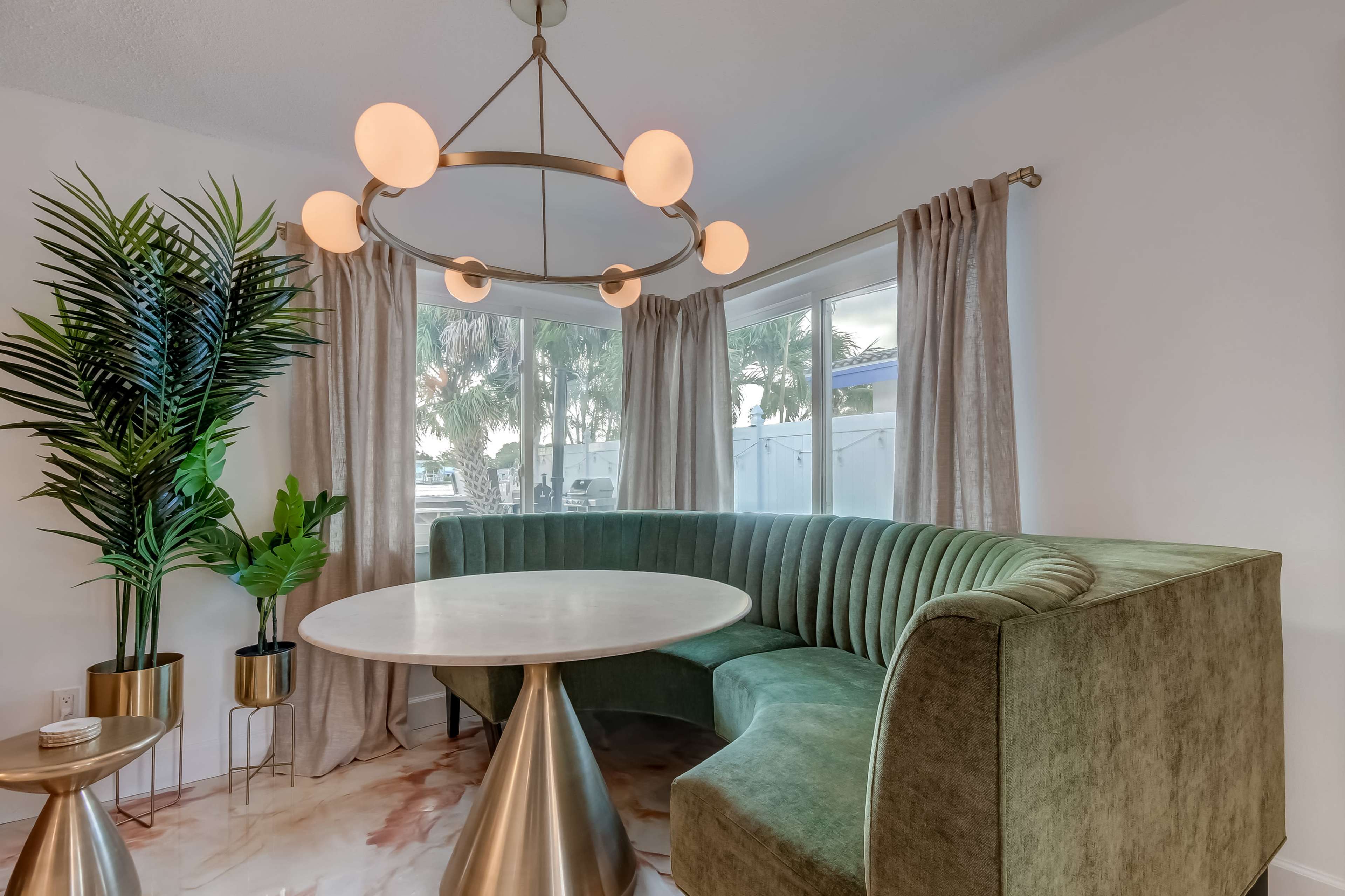 A modern dining area with a round table surrounded by a curved greenbanquette and a large chandelier, complemented by potted plants and large windows.