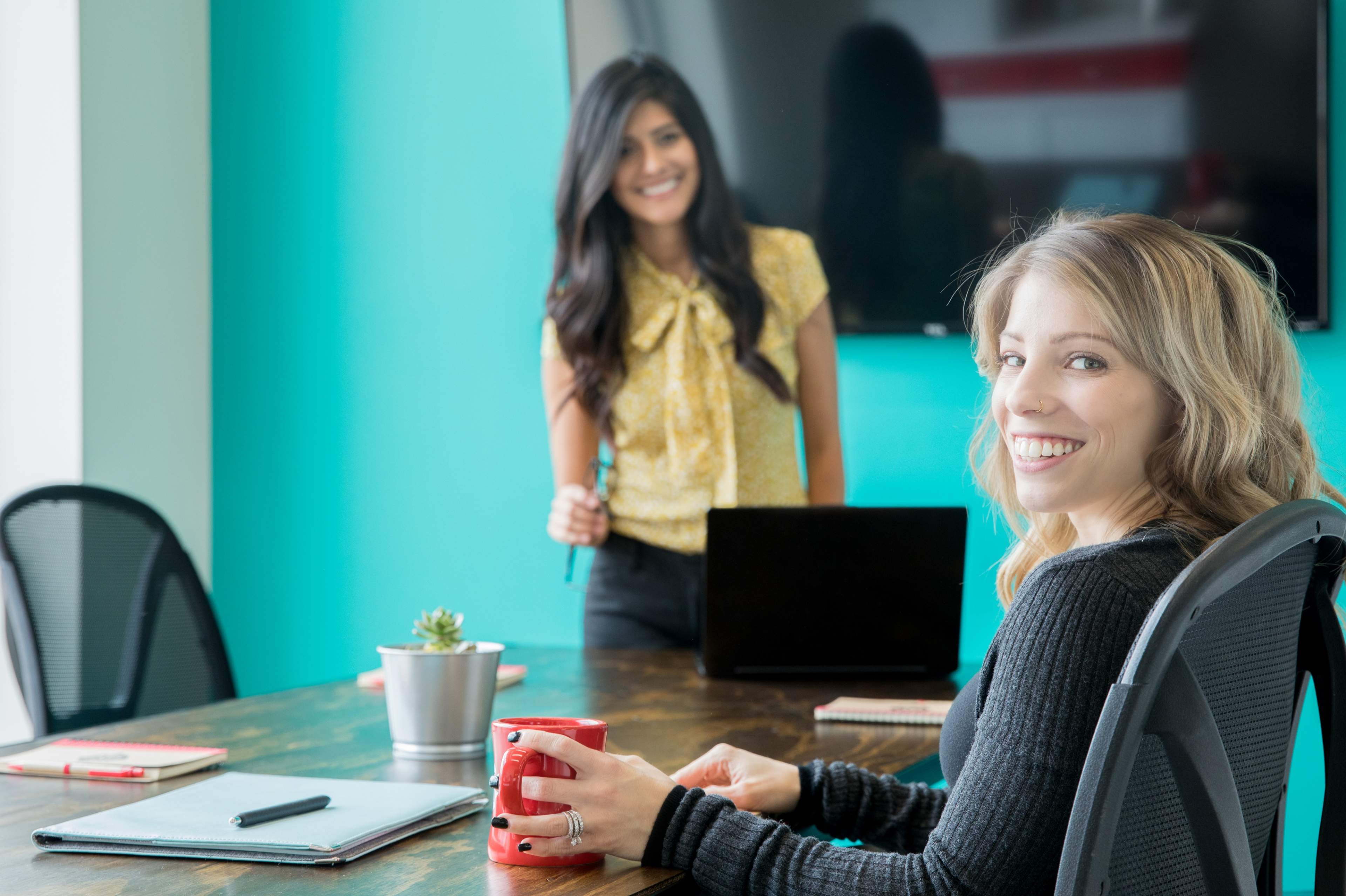 Two women are seated at a conference table, with one standing in the background and the other smiling at the camera while holding a red mug.