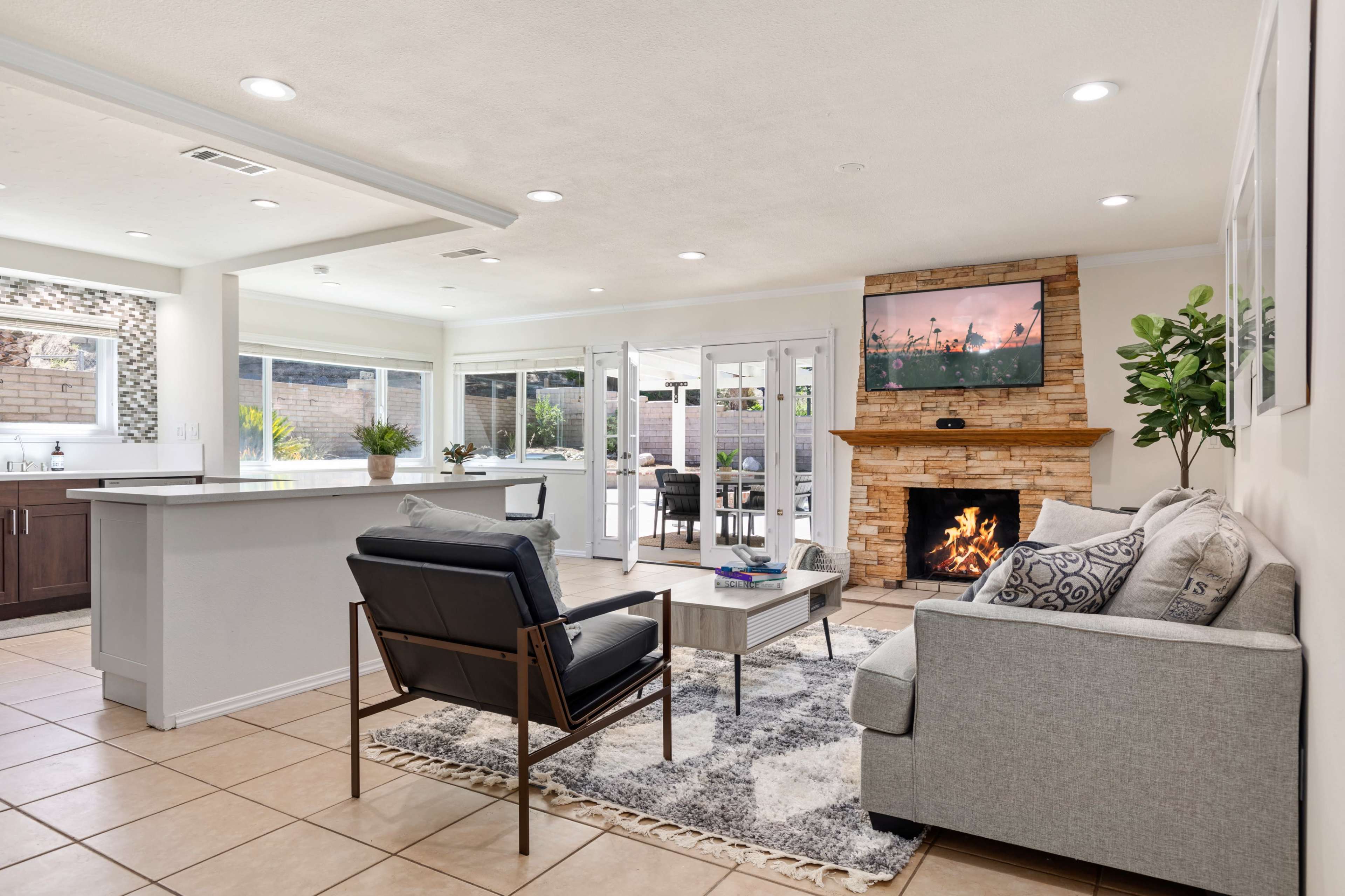 The image shows a modern living room with a stone fireplace, television mounted on the wall, and an open layout leading to an outdoor patio area.