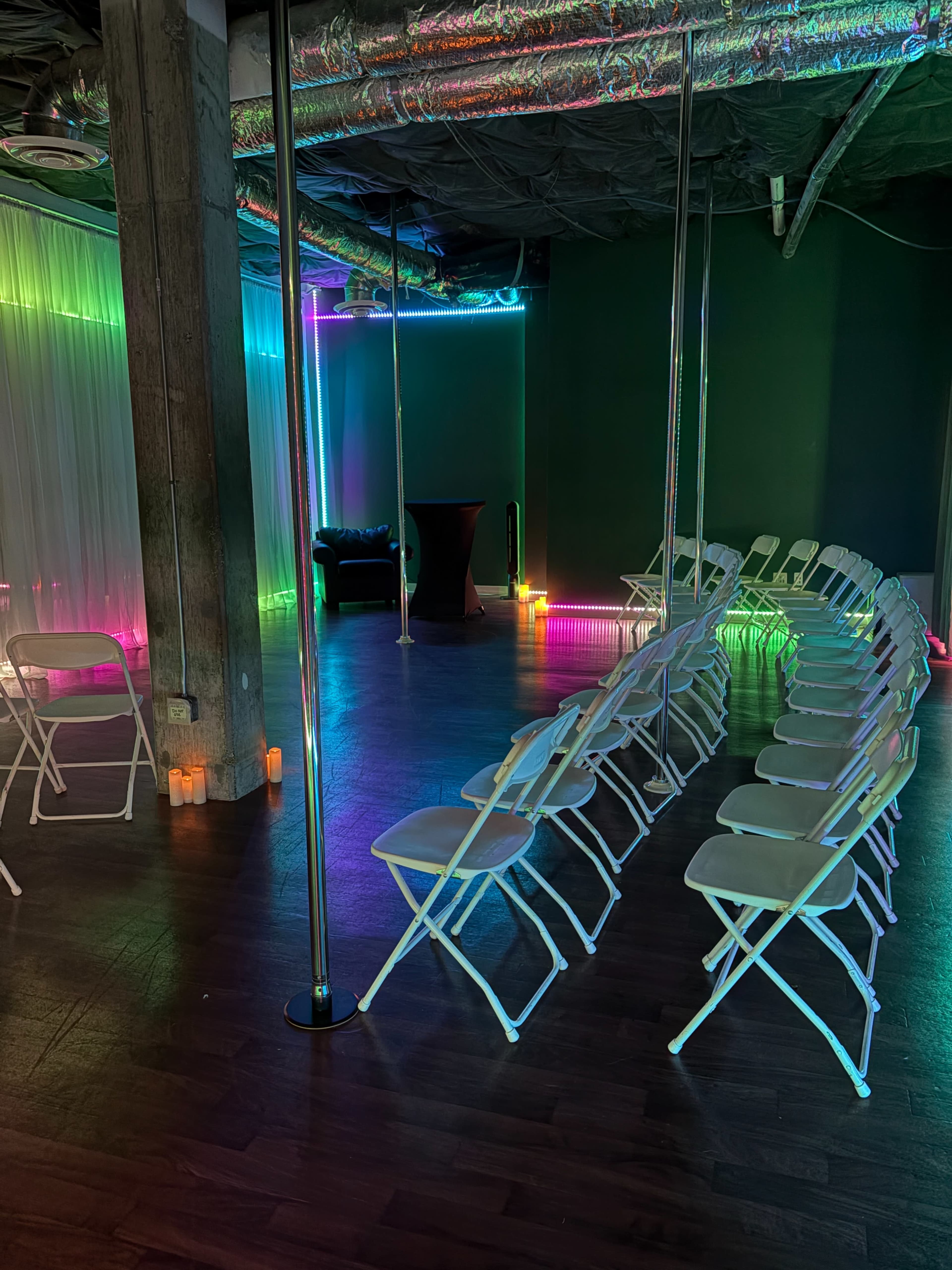 The image shows a dimly lit room set up for an event, featuring rows of white folding chairs, a pole in the center, and colorful LED lights illuminating the walls and floor.