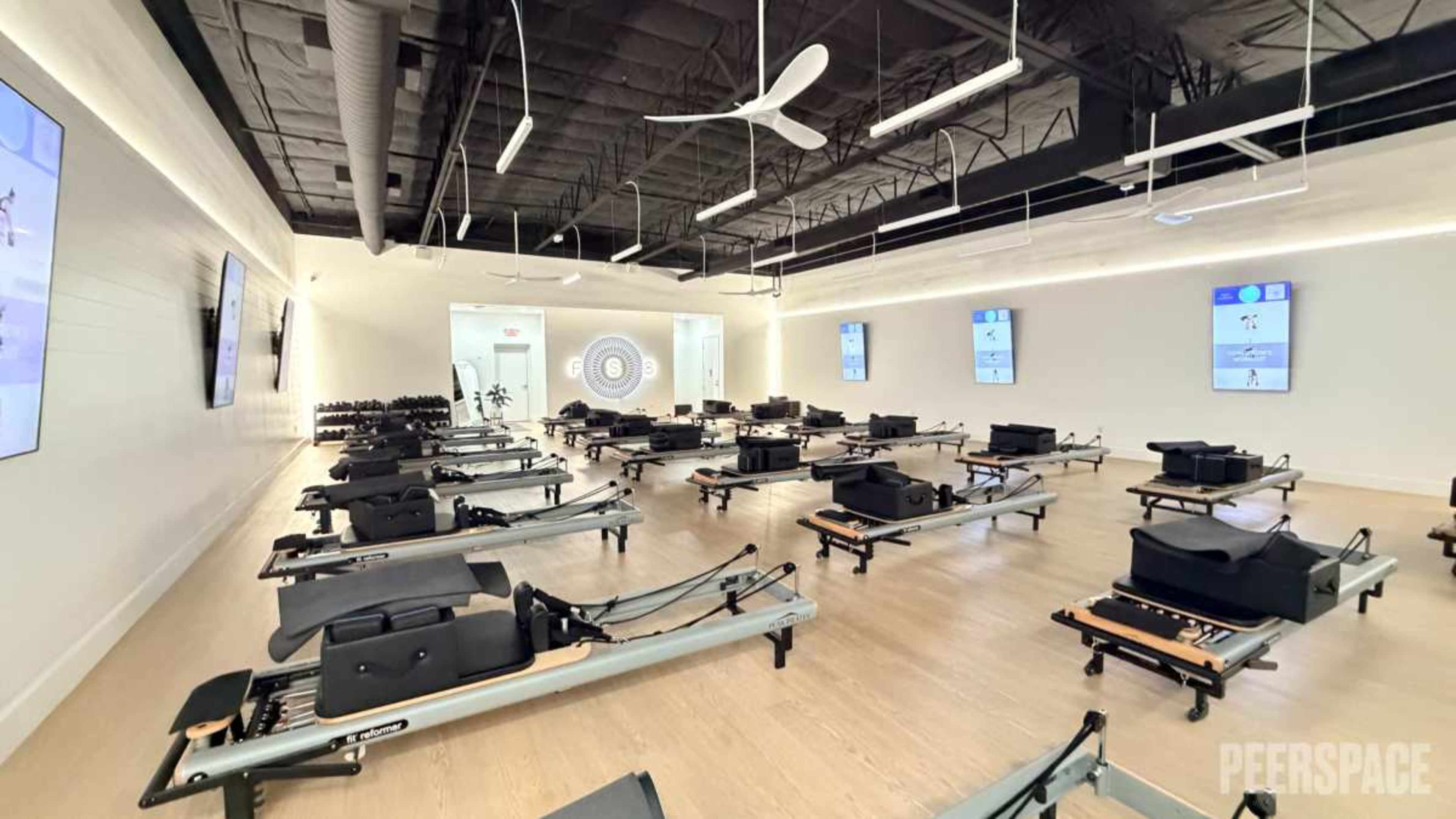 The image shows a spacious fitness studio filled with reformer Pilates machines arranged in rows, with large screens displaying workout information on the walls.