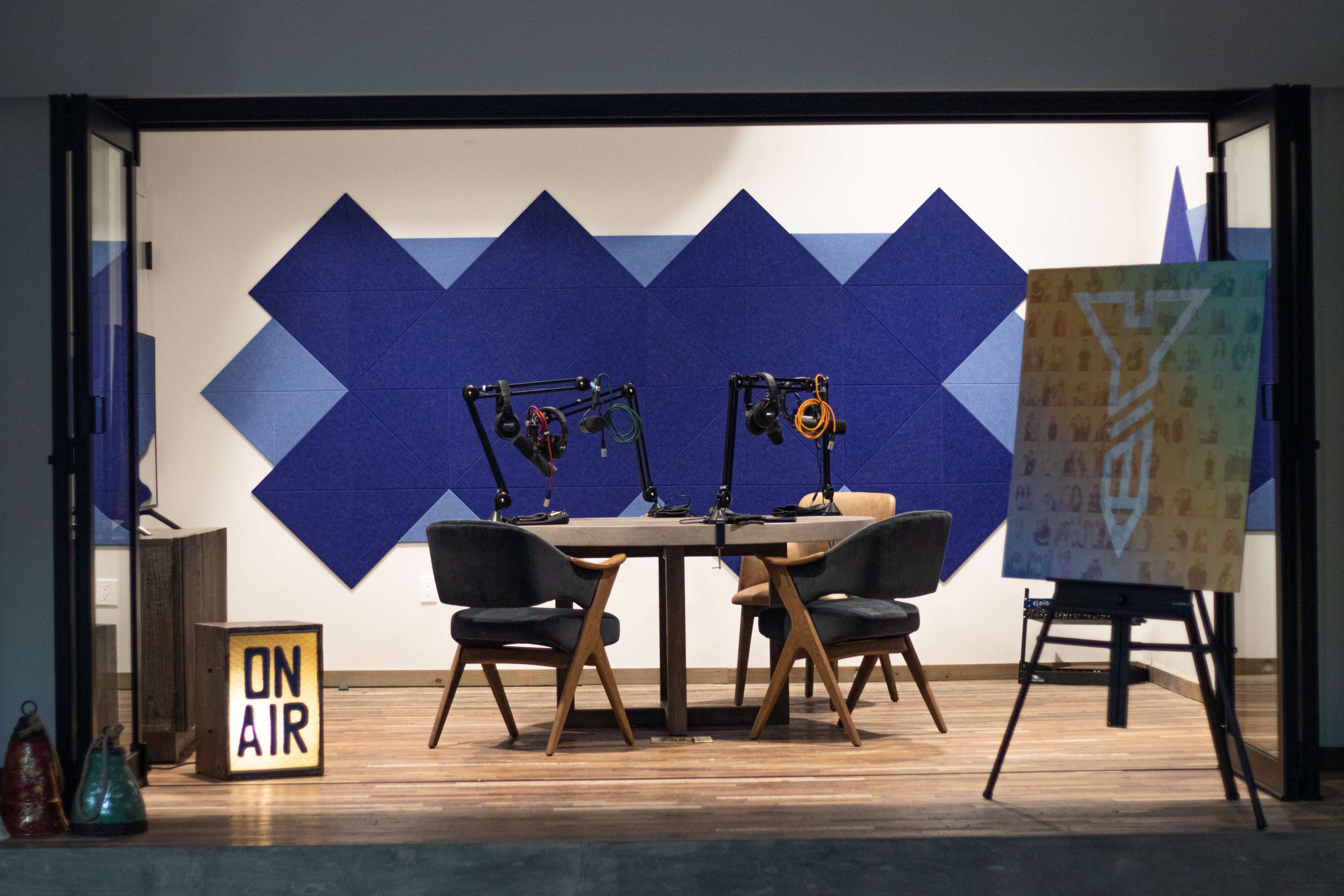 The image shows a podcast recording studio featuring a wooden table with two chairs, microphone arms, soundproof blue wall panels, and an "ON AIR" sign.