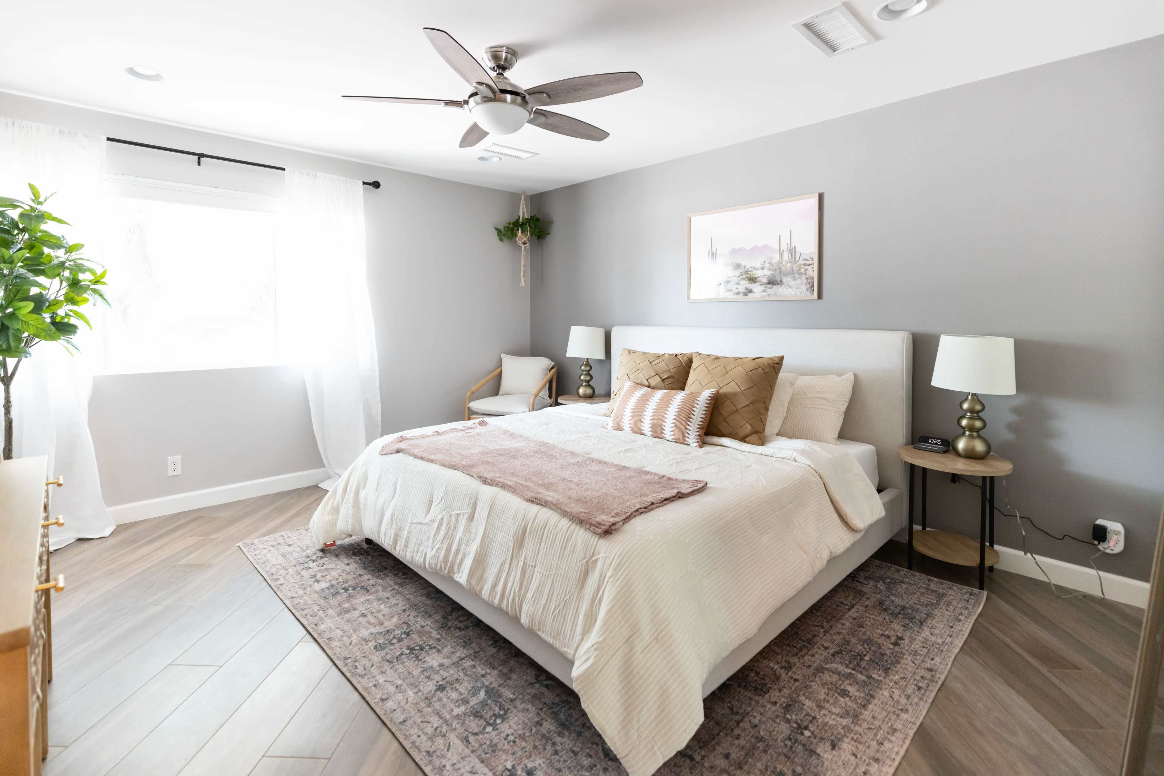 A modern bedroom features a large bed with neutral bedding, a decorative throw, and minimalistic furniture, including a chair and side tables with lamps.