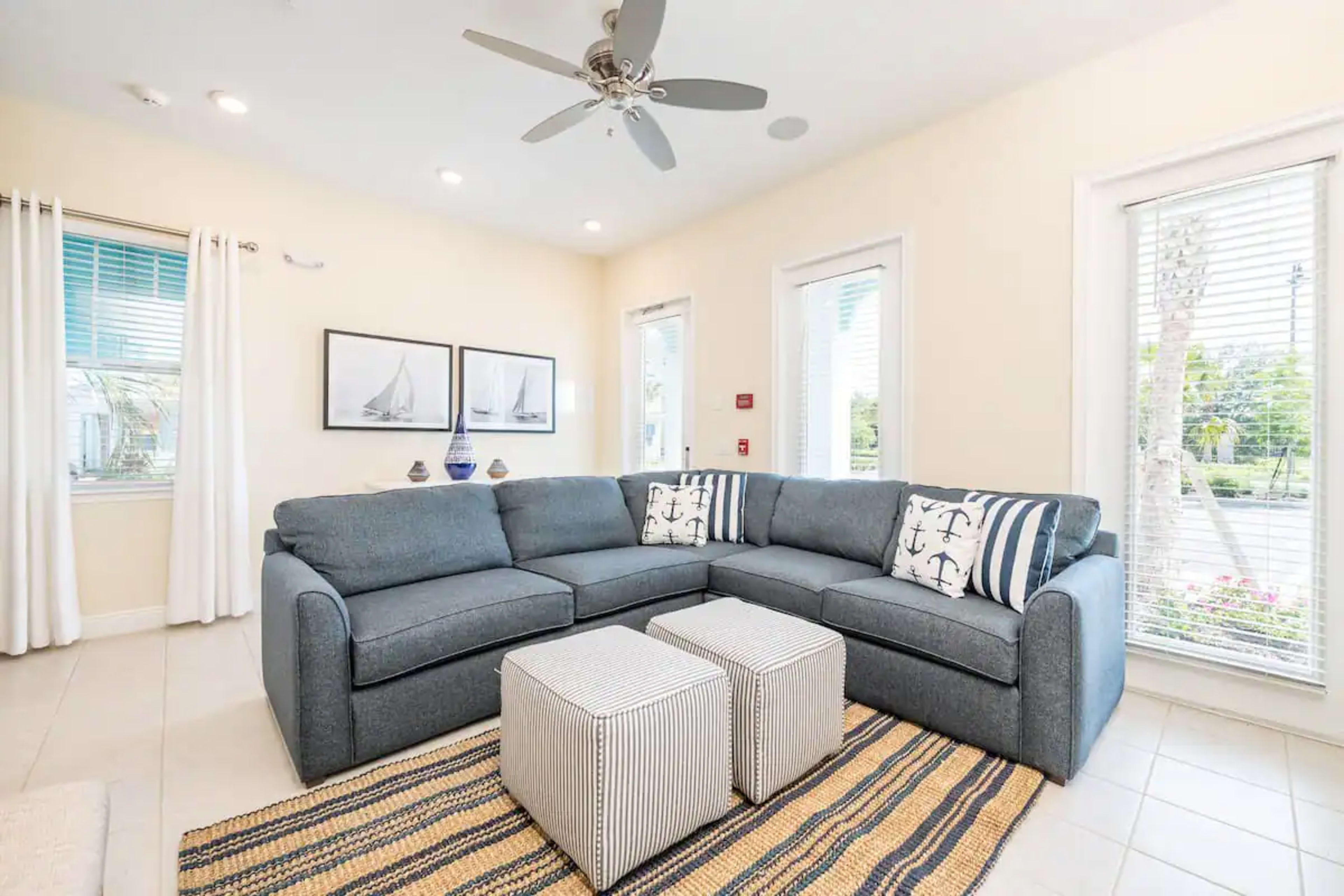 The image shows a cozy living room with a sectional sofa, two square ottomans, and large windows allowing natural light to enter.