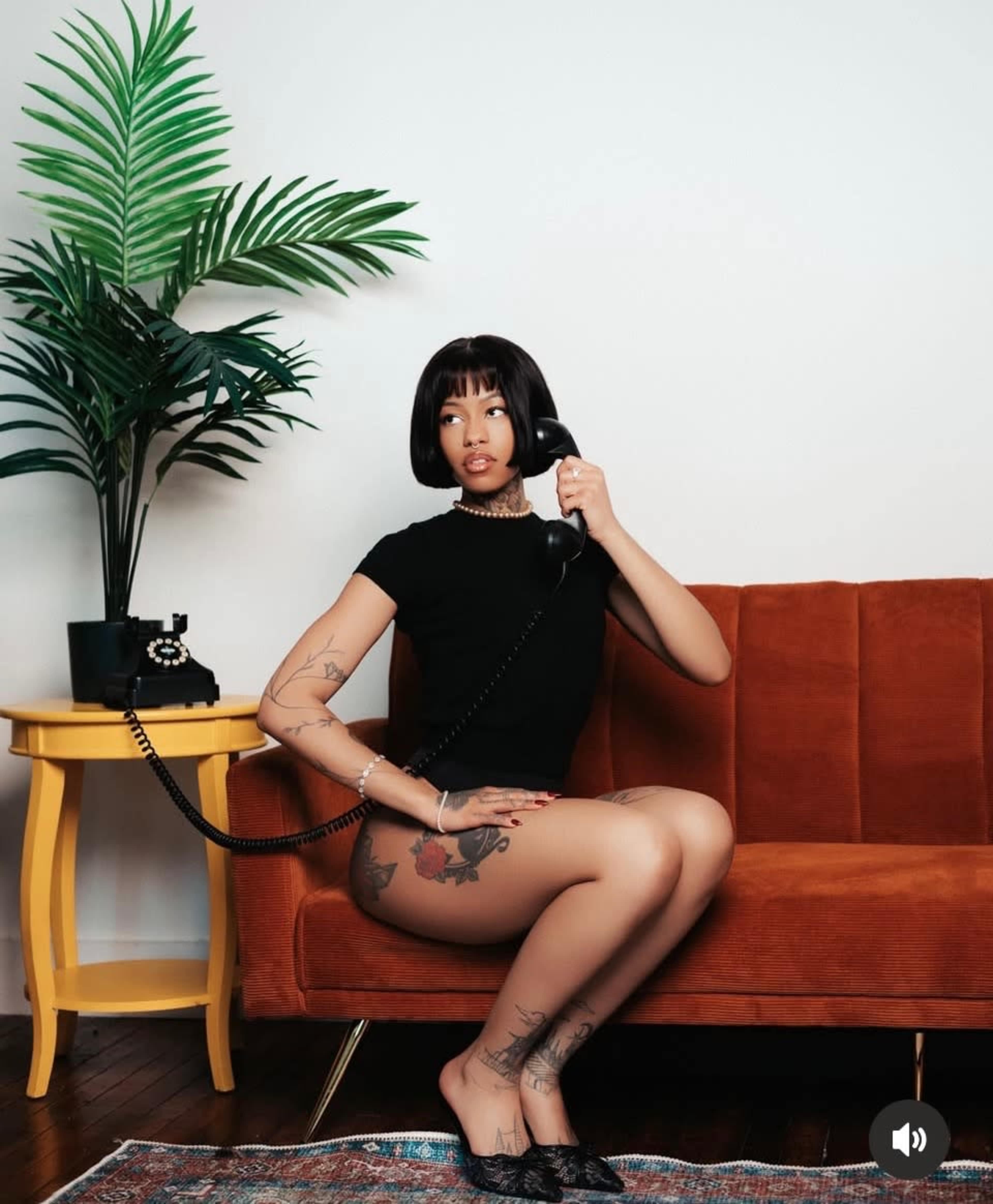 A person sits on a brown couch while holding a vintage telephone, with a potted plant and a yellow side table in the background.