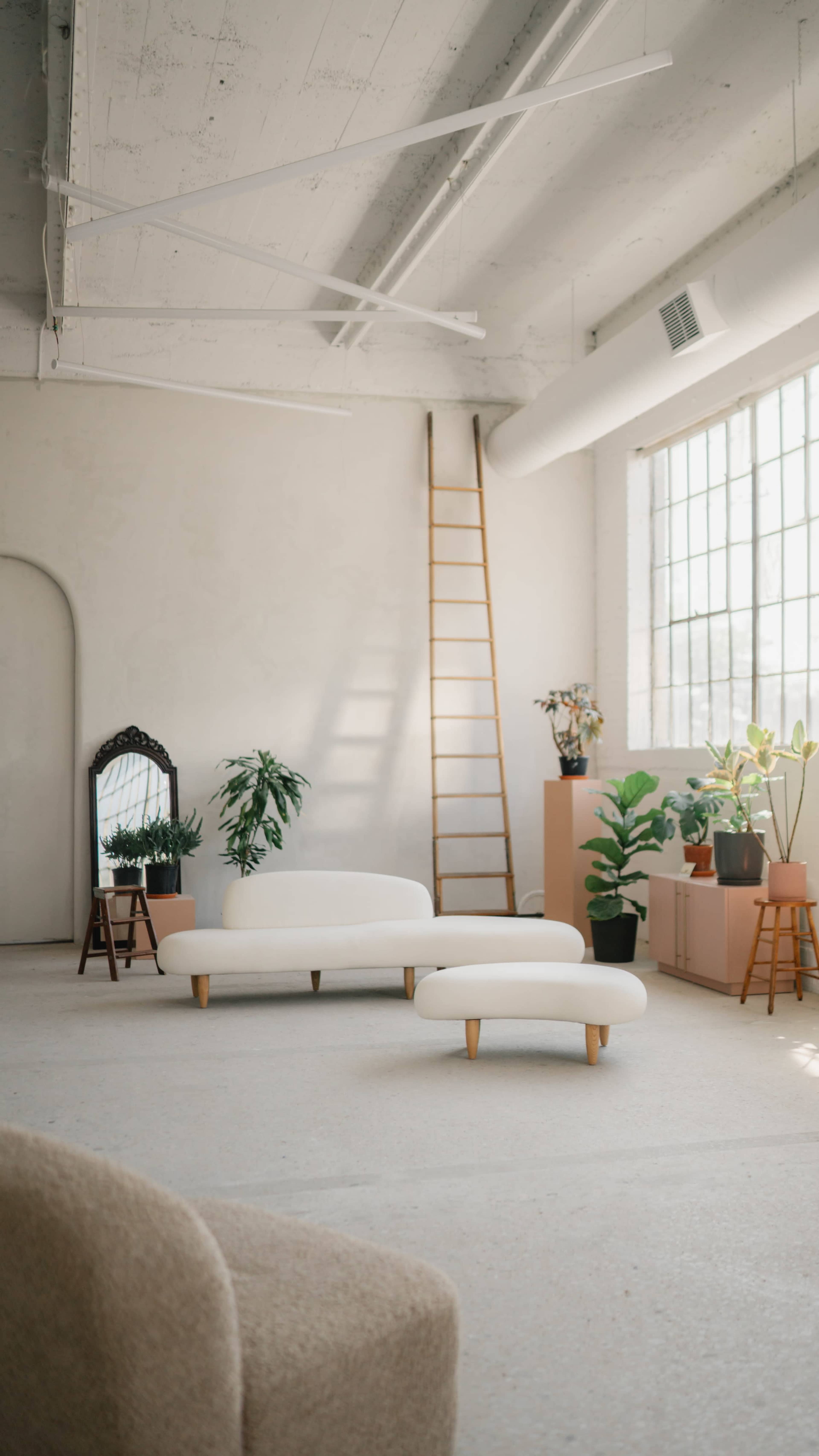 A bright, minimalist interior featuring a white sofa, a smaller white coffee table, plants, a ladder against the wall, a mirror, and a window allowing natural light to fill the space.