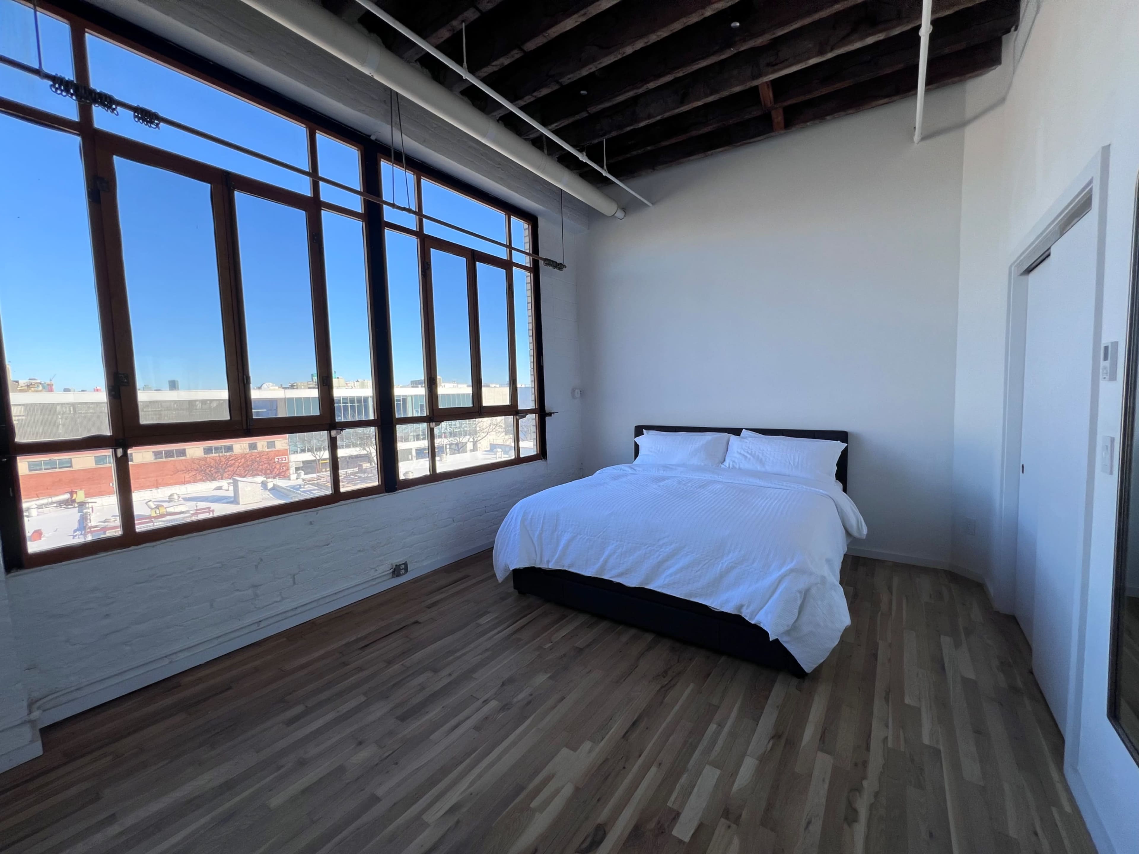 The image shows a bright, minimalistic bedroom featuring a bed against a large window, with hardwood flooring and exposed beams in the ceiling.