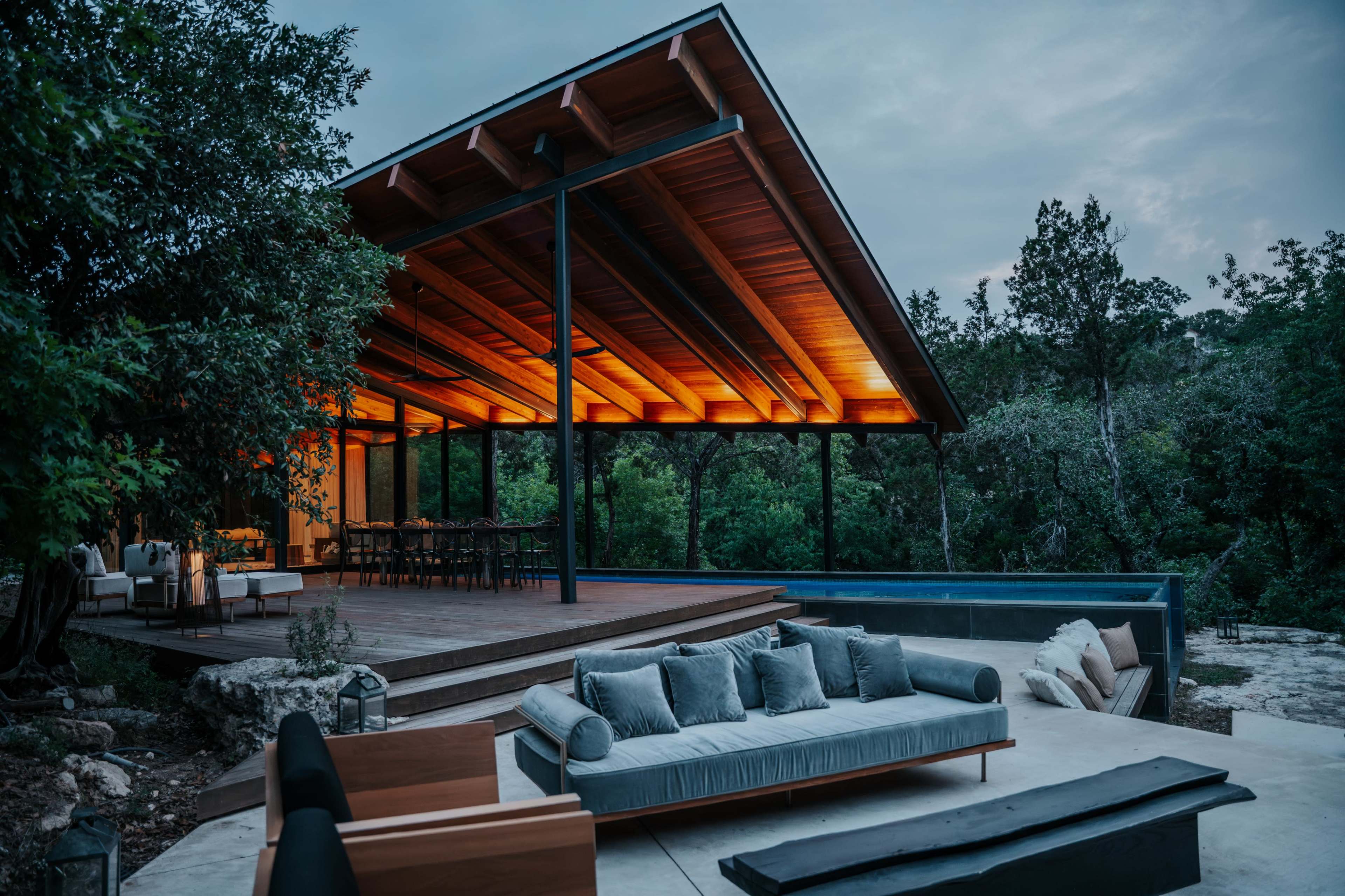 A modern outdoor living space features a wooden deck with a sleek sofa and a dining area under an expansive roof, surrounded by greenery.