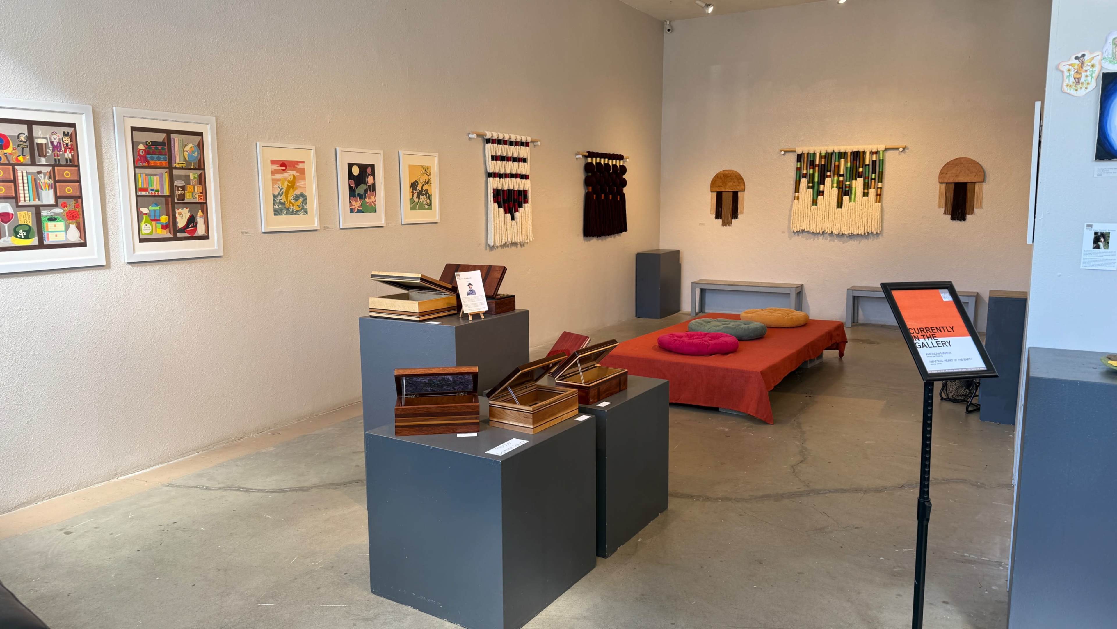The image depicts an art gallery showcasing various wooden boxes on display stands, colorful circular cushions, and framed artworks on the walls.