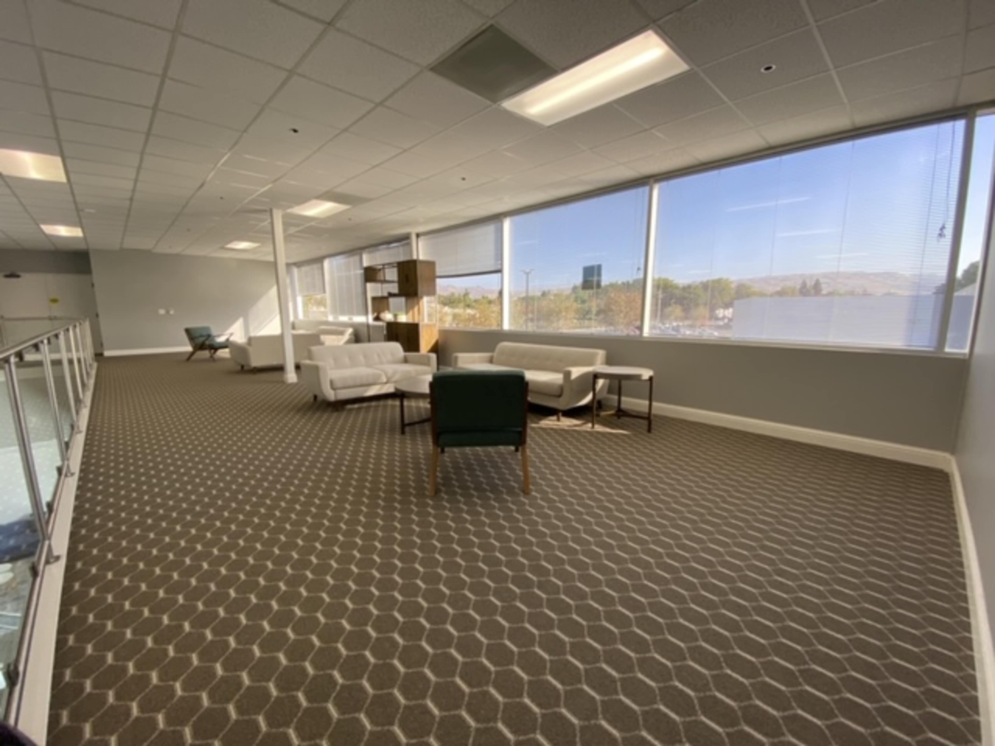 The image shows an empty office space with white sofas, a green chair, and large windows allowing natural light to enter.