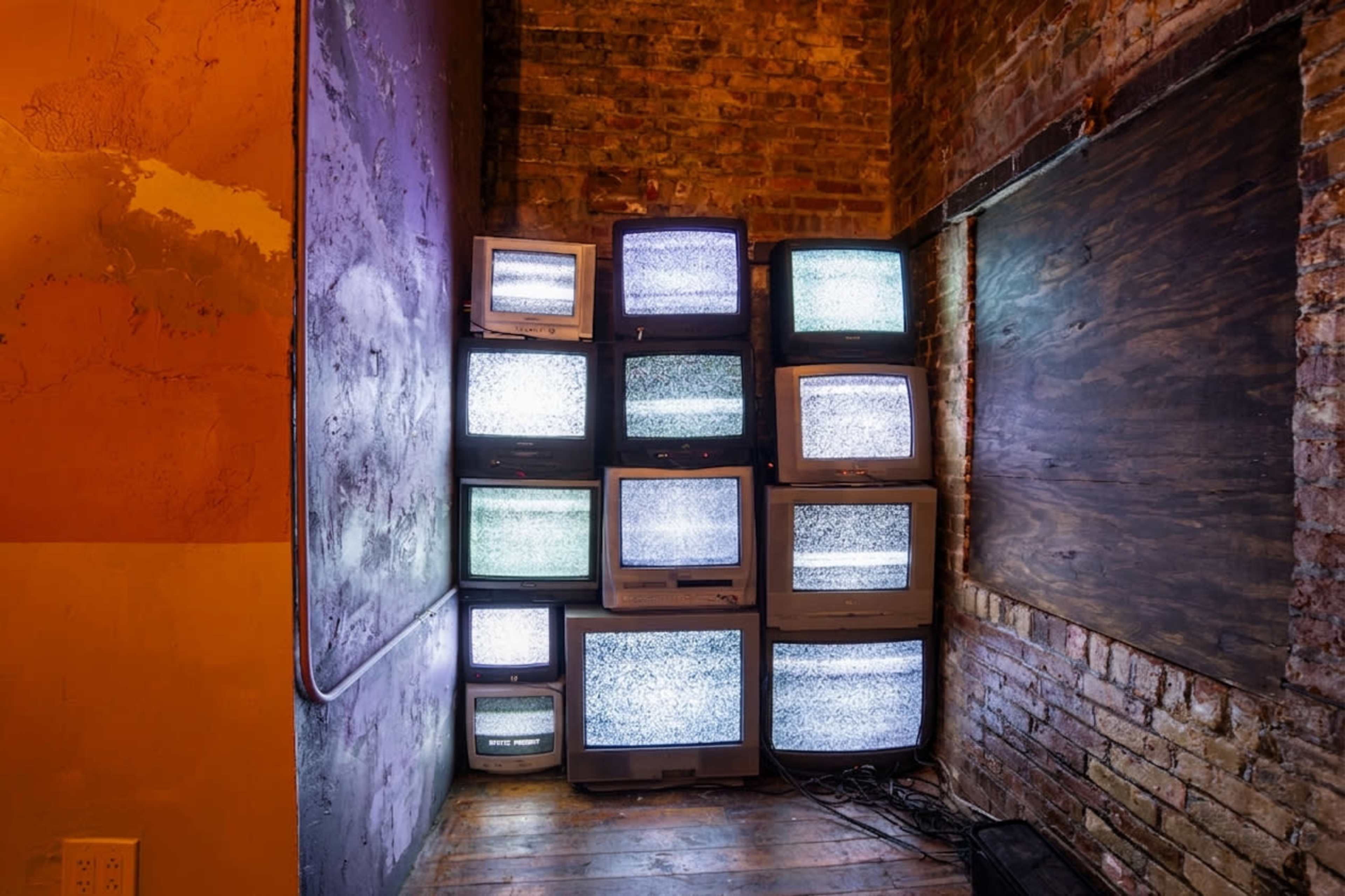 A stack of old television sets displaying static is positioned against a brick wall in a dimly lit room.