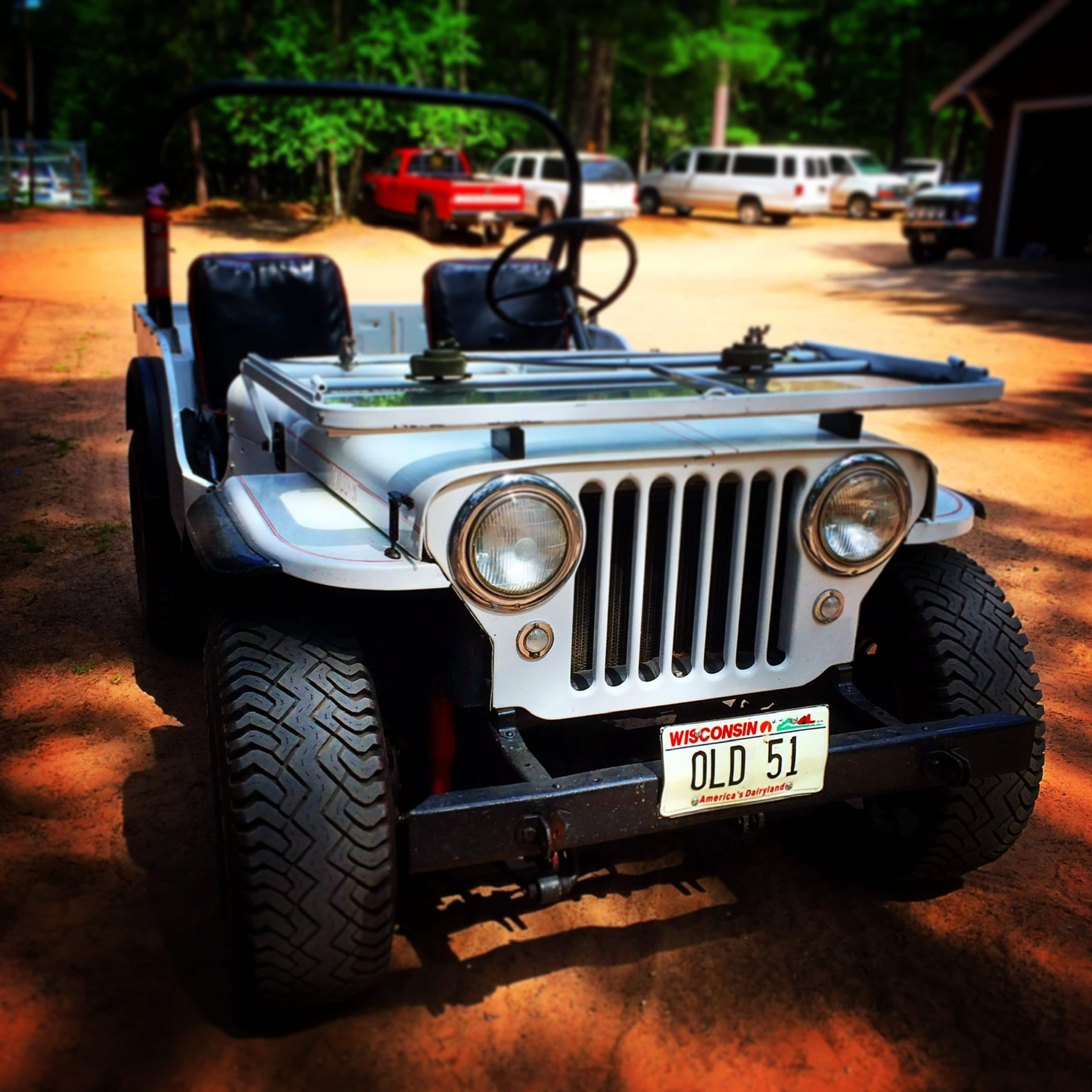 A vintage Jeep with a Wisconsin license plate reading "OLD 51" is parked on a dirt driveway surrounded by trees and other vehicles in the background.