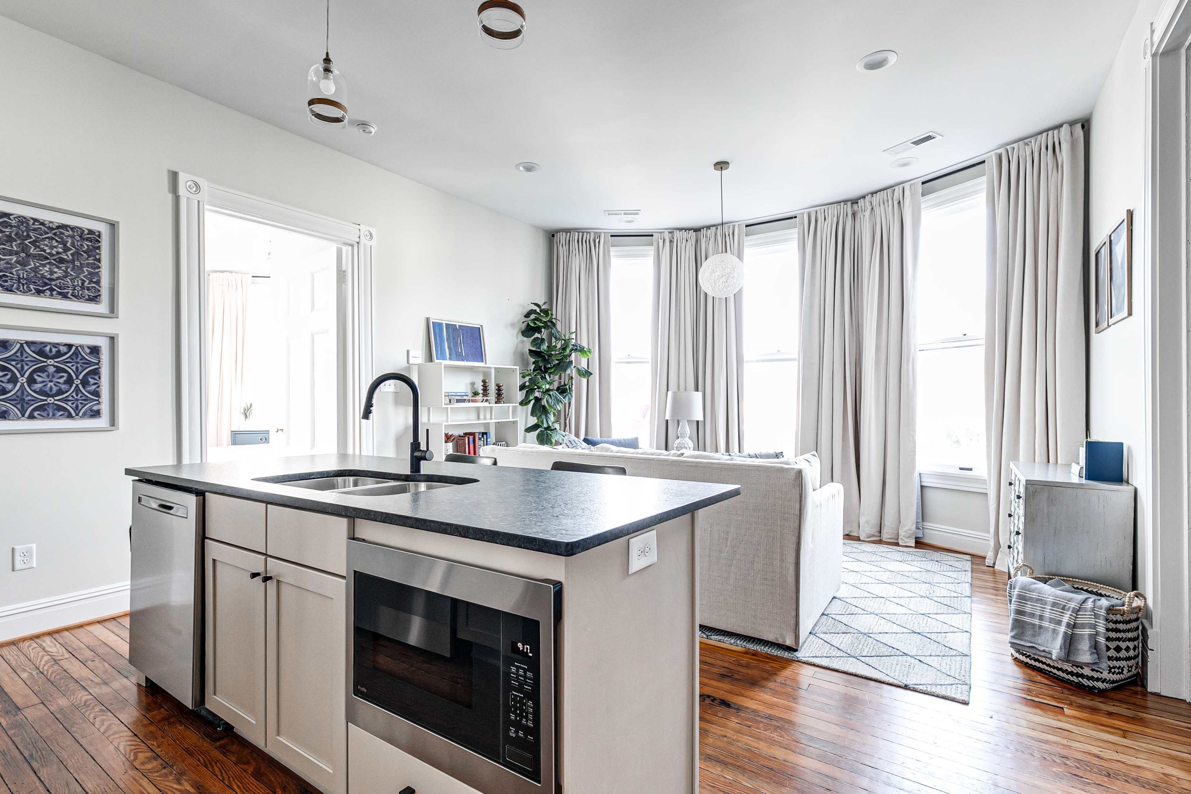 A modern kitchen features a central island with a microwave, overlooking a bright living area with large windows and light-colored curtains.