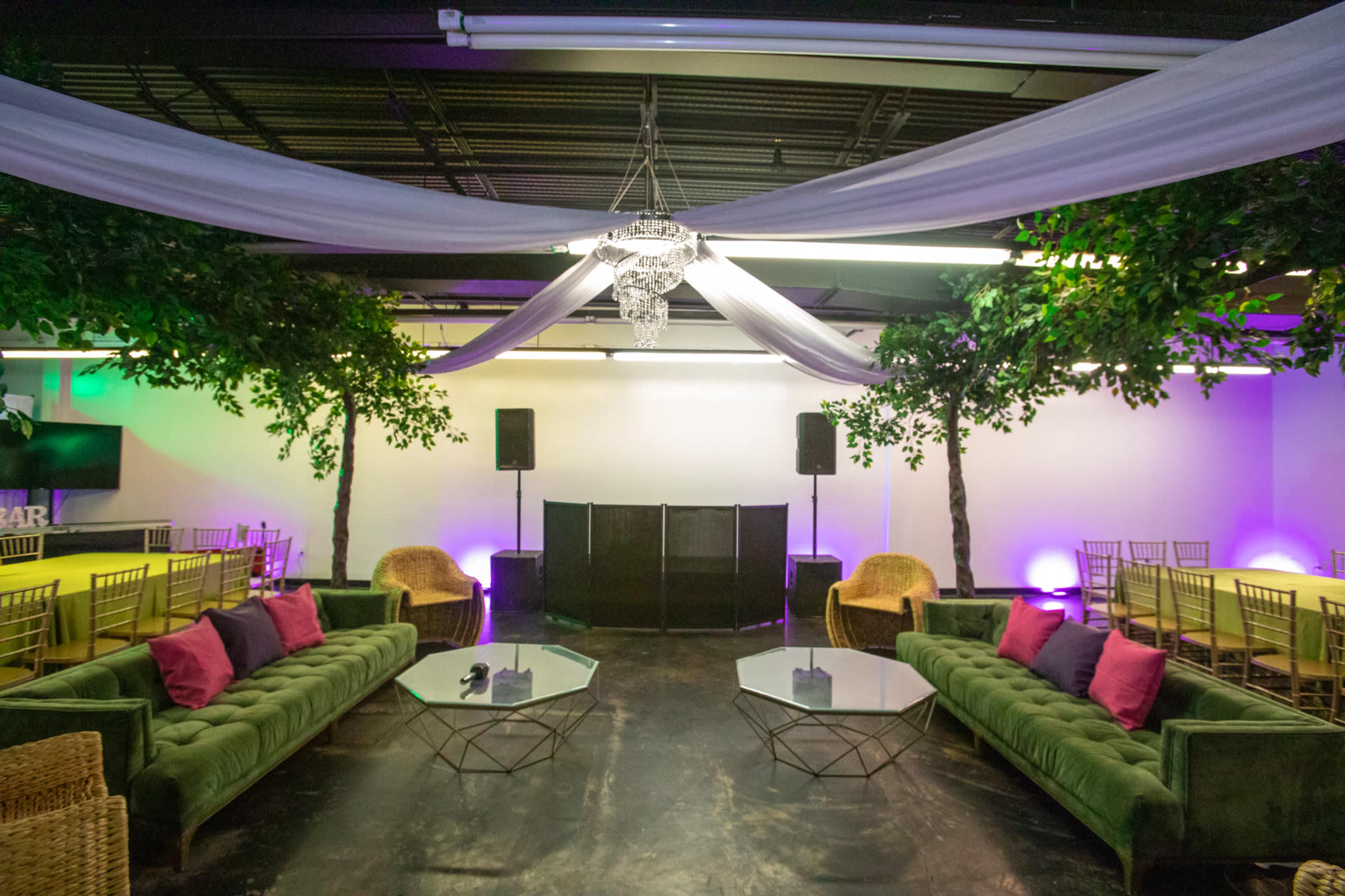 A spacious event venue with green sofas, glass coffee tables, and a stage backdrop adorned with trees and draped fabric.