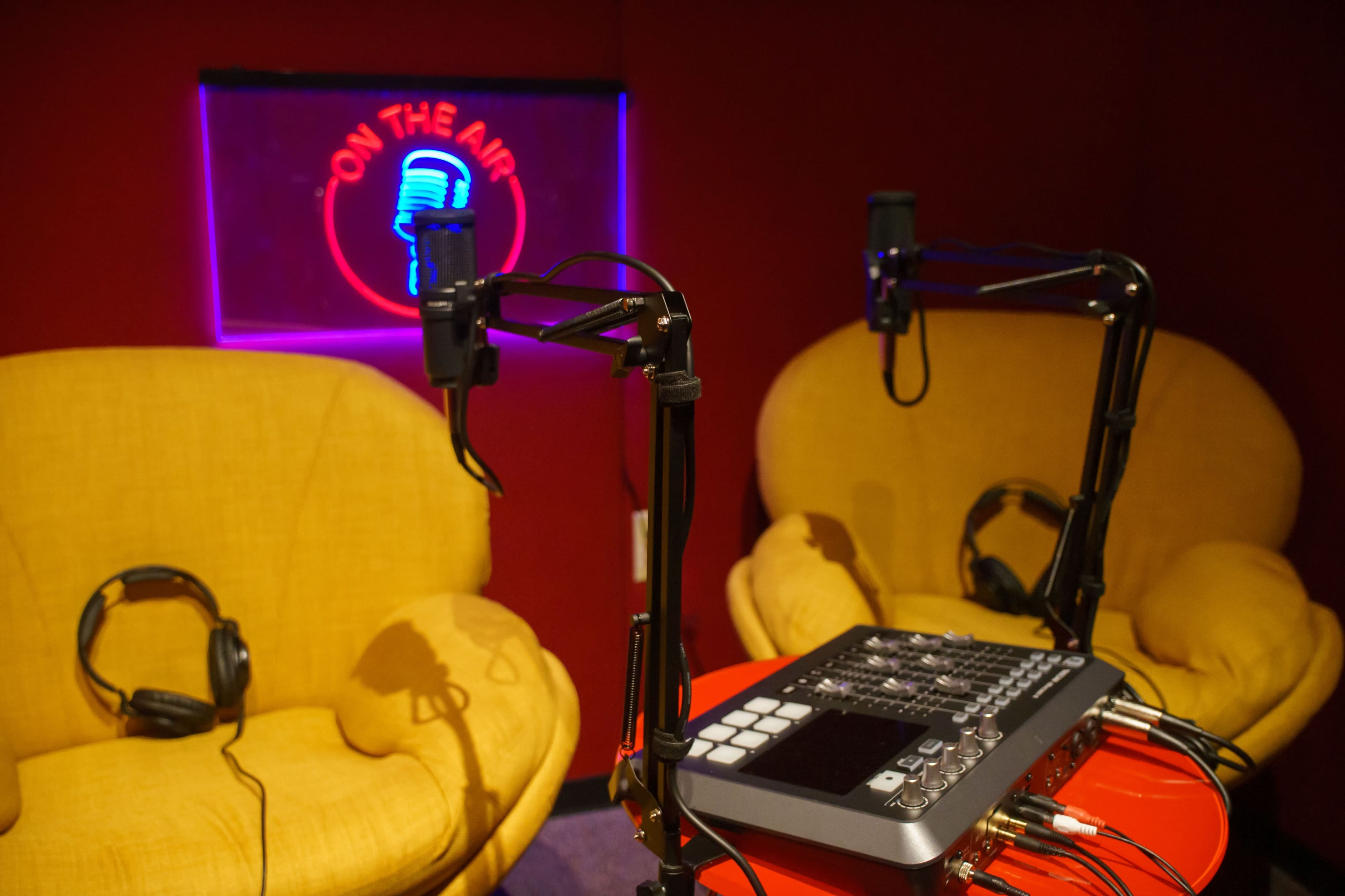 The image shows a radio studio setup with two yellow armchairs, a sound mixer on a red table, and a neon sign that reads "ON THE AIR."