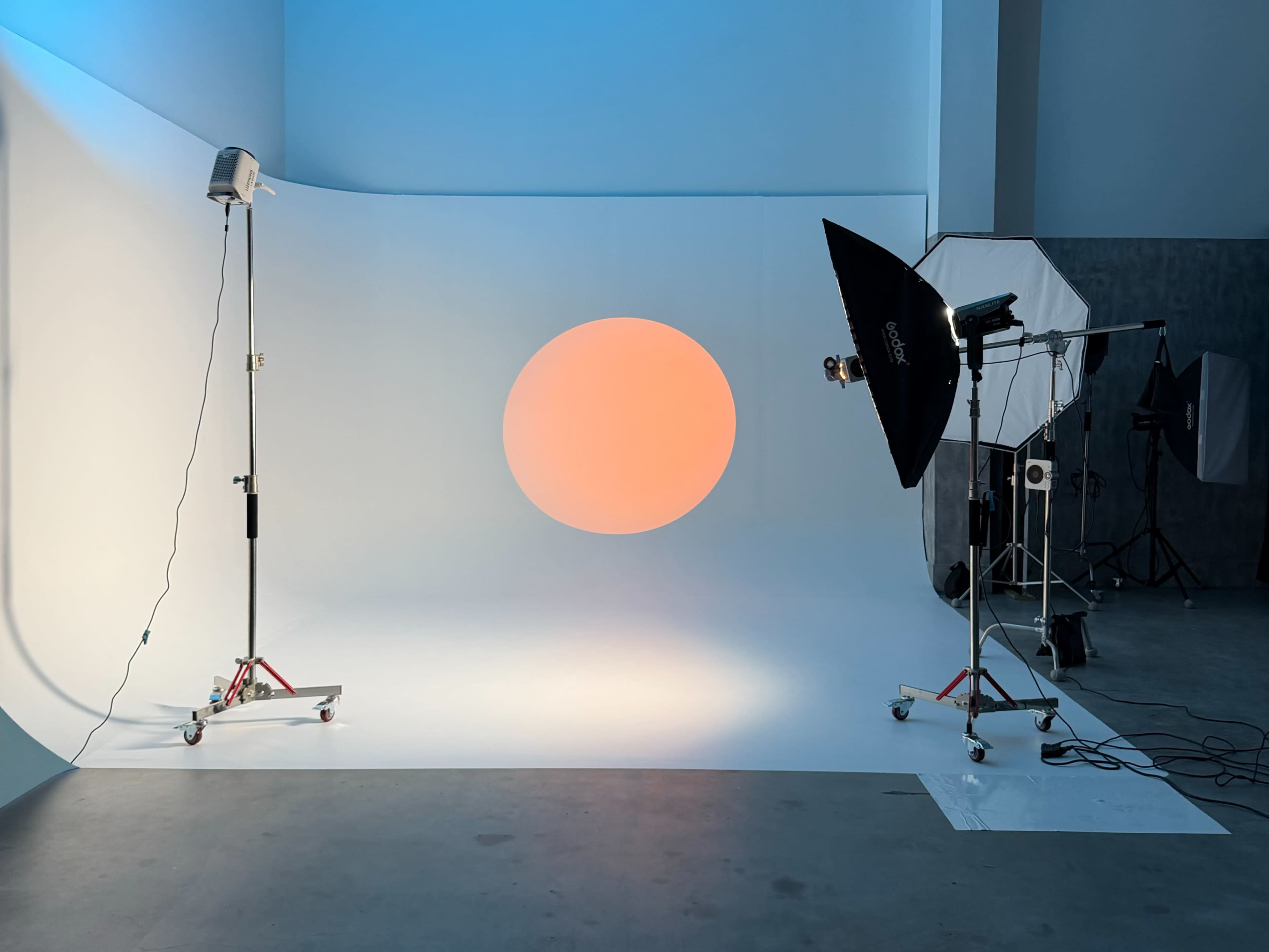 A photo studio setup features a white backdrop with an orange circular light, flanked by a softbox and a light stand.
