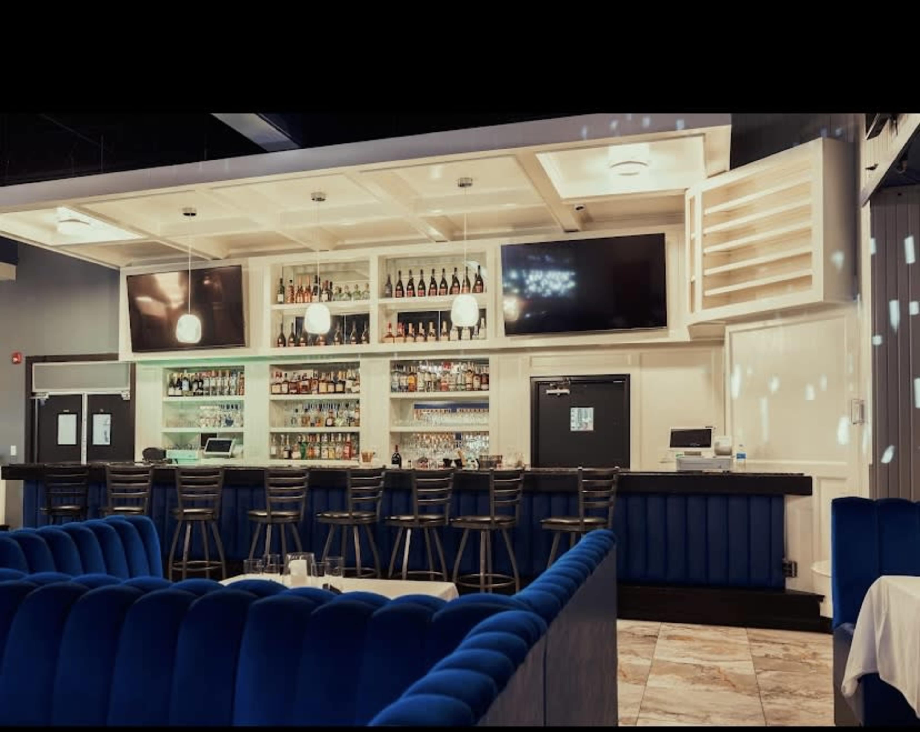 The image features a modern bar area with a well-stocked liquor shelf, multiple TV screens, and seating at a blue velvet-accented counter.
