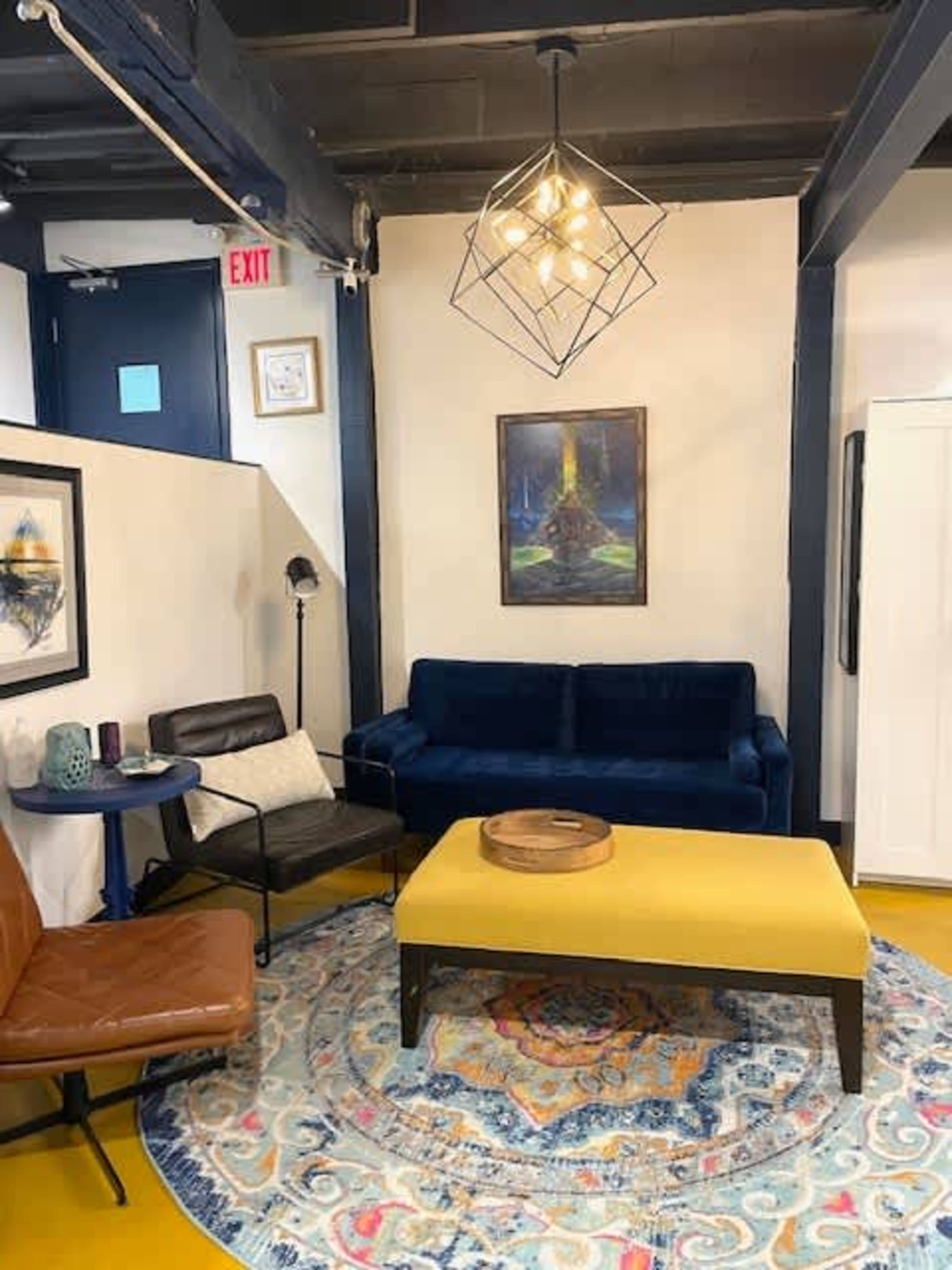 The image shows a small, modern sitting area with a dark blue sofa, a yellow coffee table, an area rug, and framed artwork on the walls.