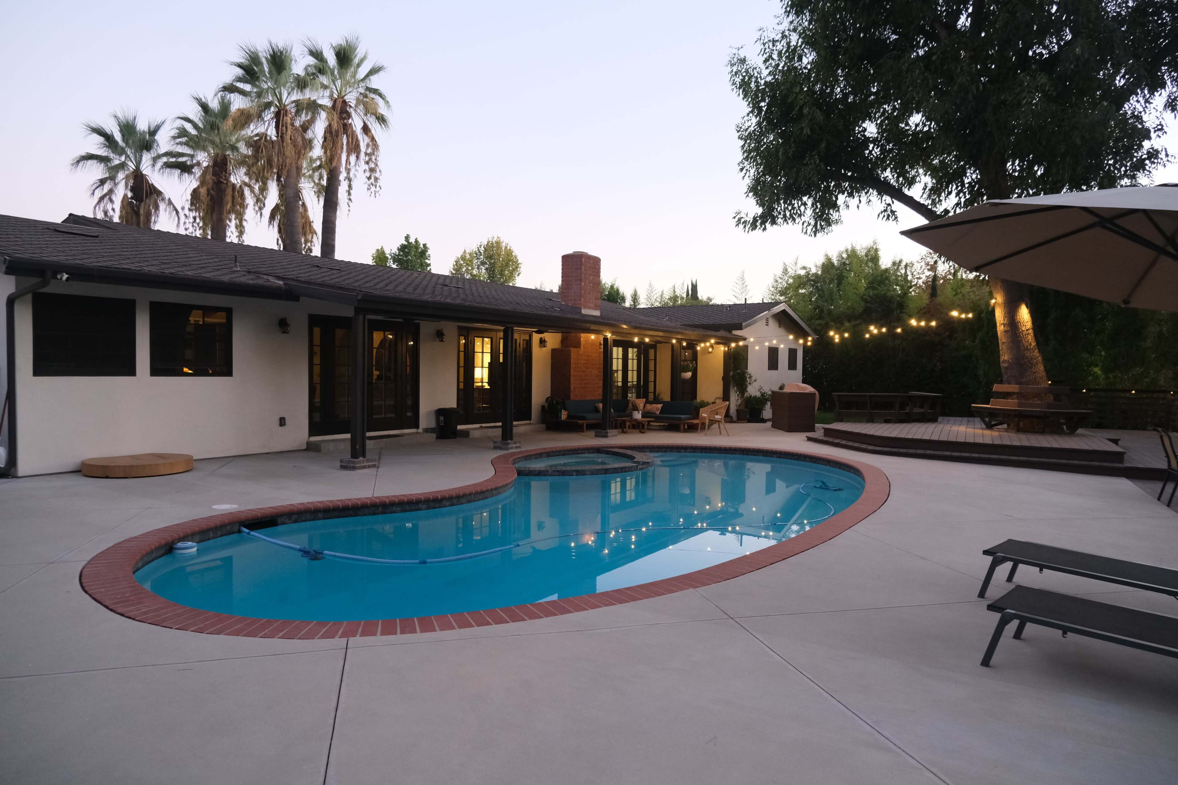 A swimming pool is situated in a backyard surrounded by palm trees and outdoor seating areas, with twilight lighting and a wooden deck nearby.