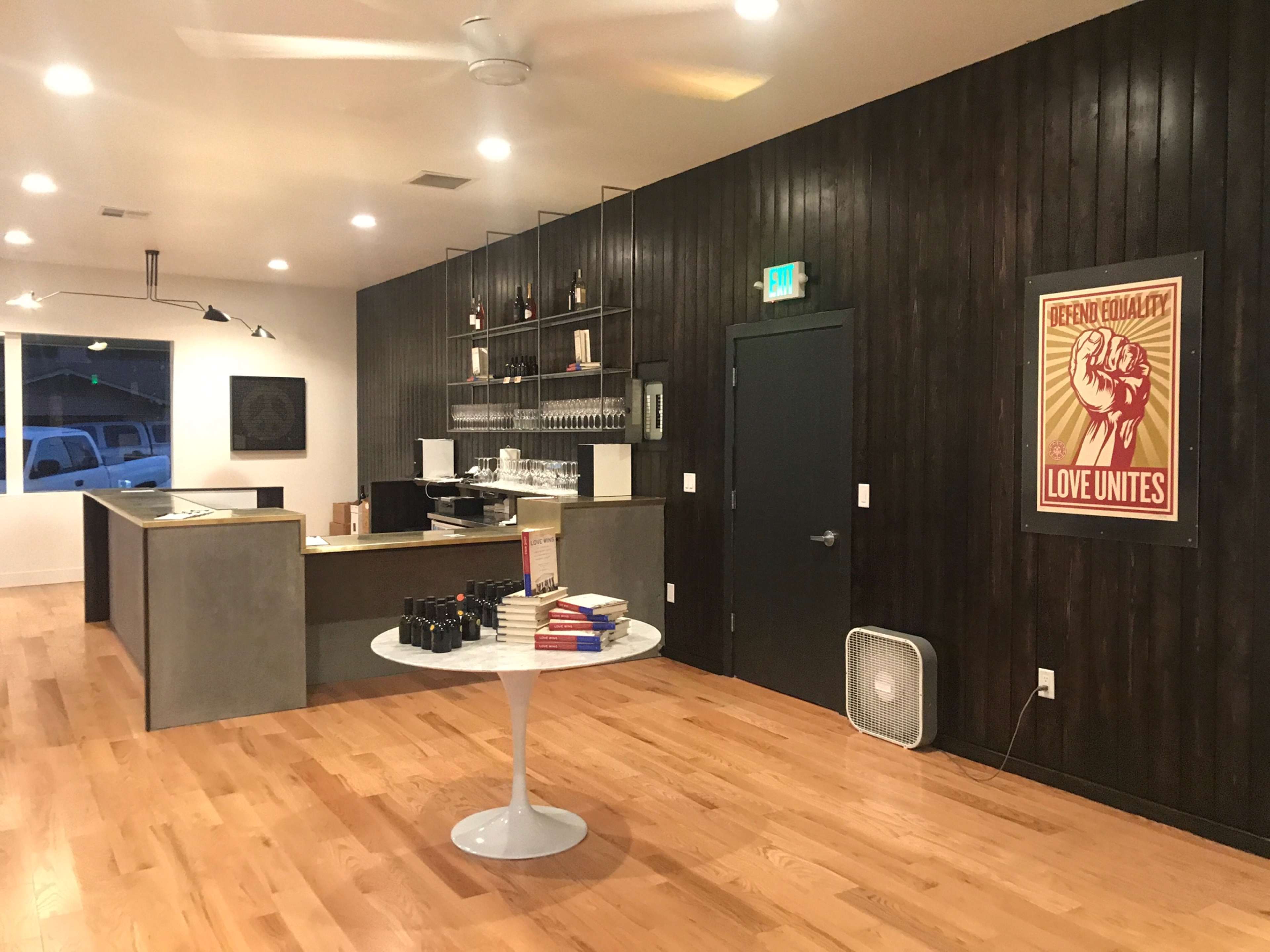 The interior features a modern, open space with a wooden accent wall, minimalist bar area, and a circular table displaying wine bottles and books.