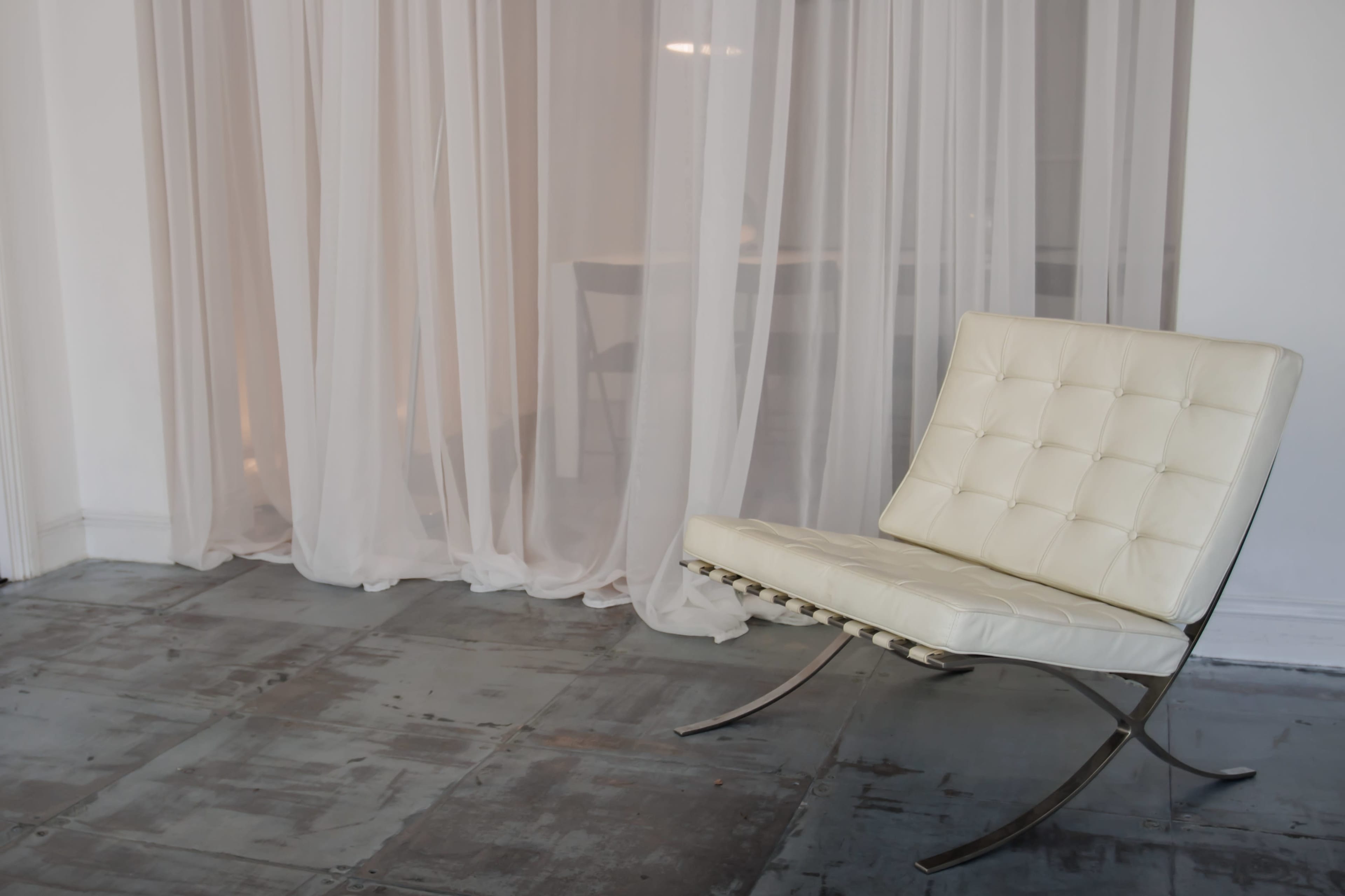 A white tufted chair sits beside sheer white curtains in a minimalistic room with a concrete floor.