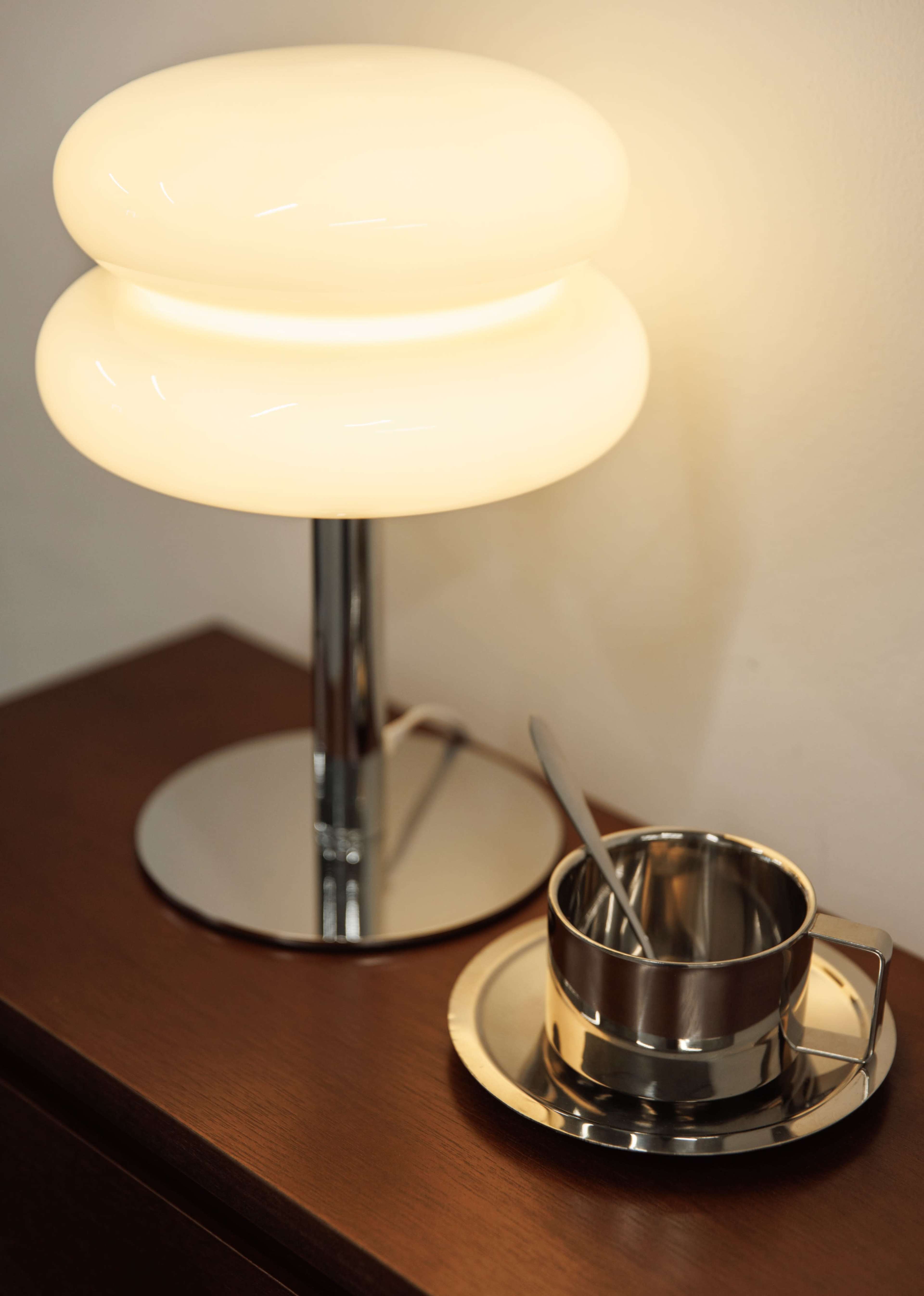 A modern lamp with a rounded glass shade stands beside a metal cup and saucer on a wooden surface.
