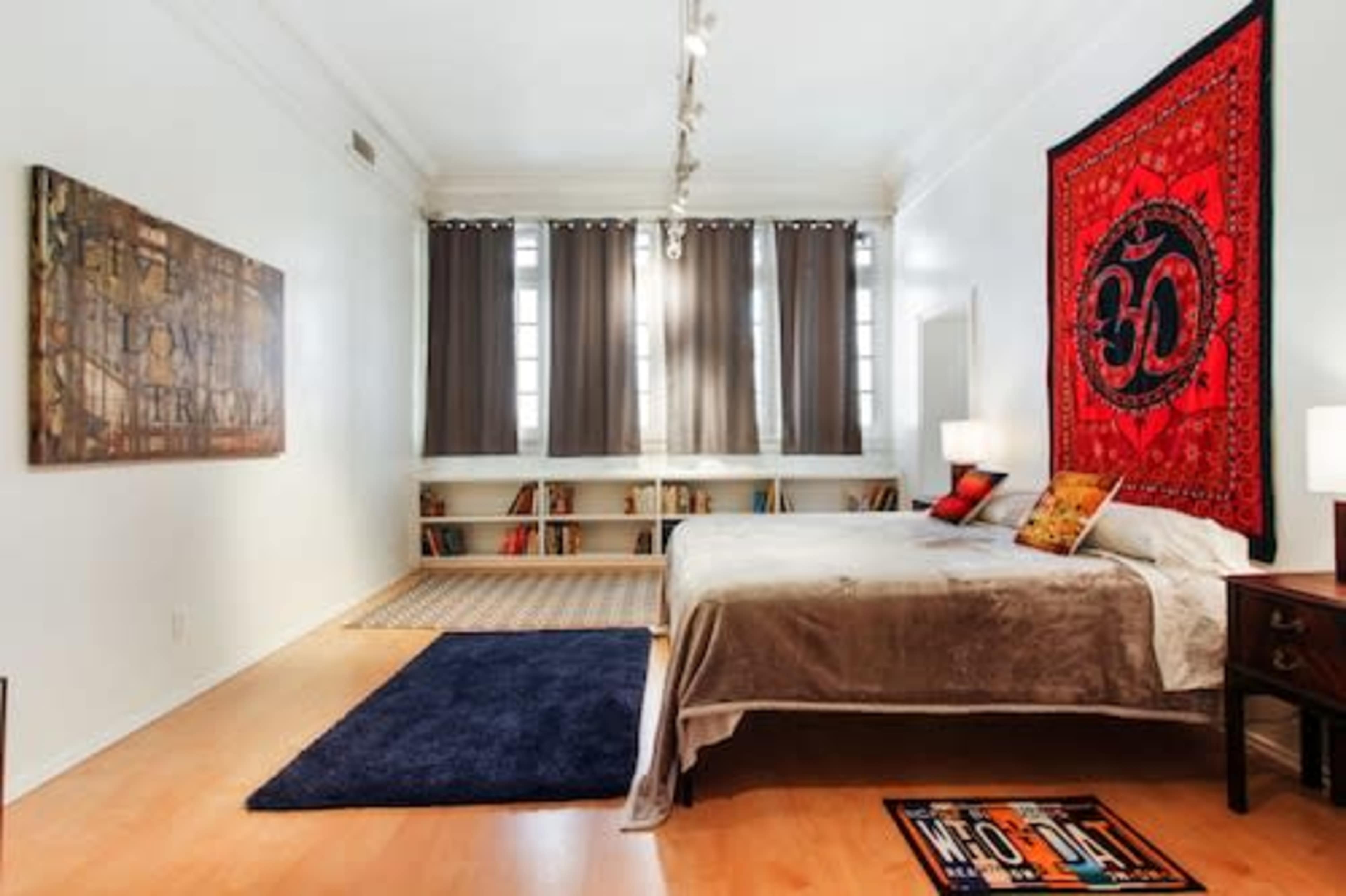 The image shows a bedroom with a bed, two bedside lamps, a red wall tapestry featuring an "Om" symbol, and bookshelves in the background.