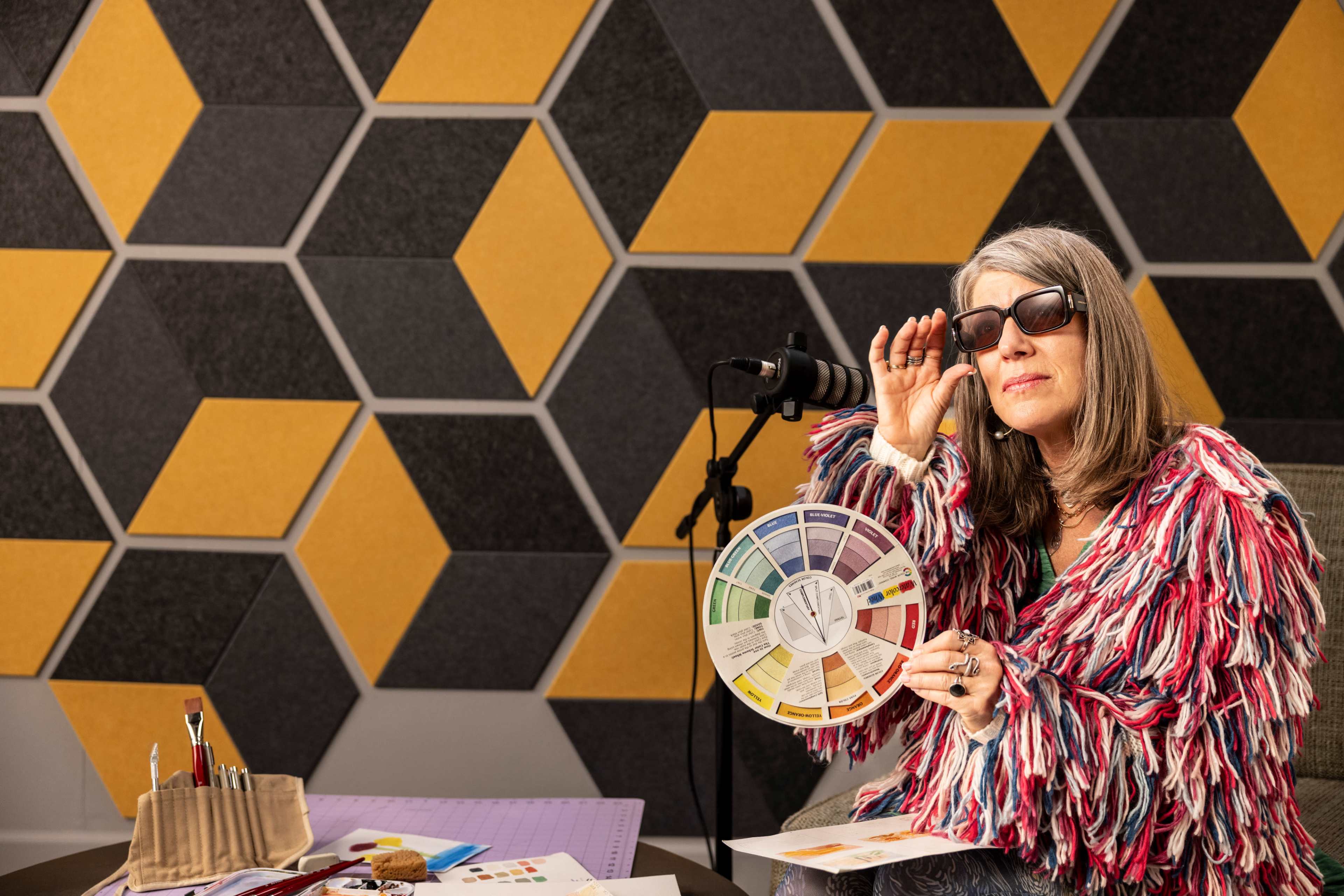 A woman wearing sunglasses holds a color wheel while sitting in front of a geometric patterned wall featuring yellow and gray hexagons.