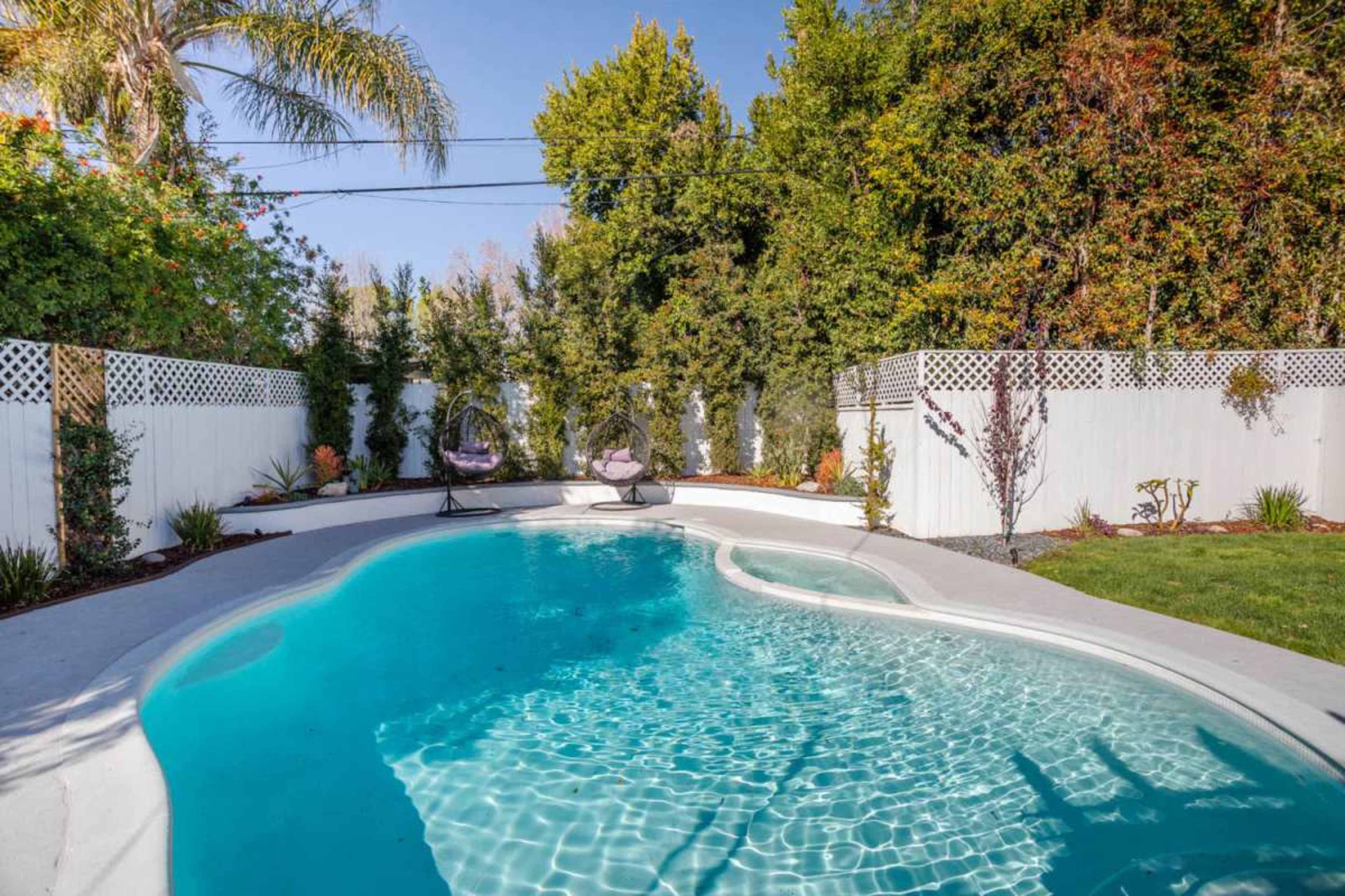 The image shows a swimming pool surrounded by greenery and a white fence, with two loungers positioned near the water's edge.