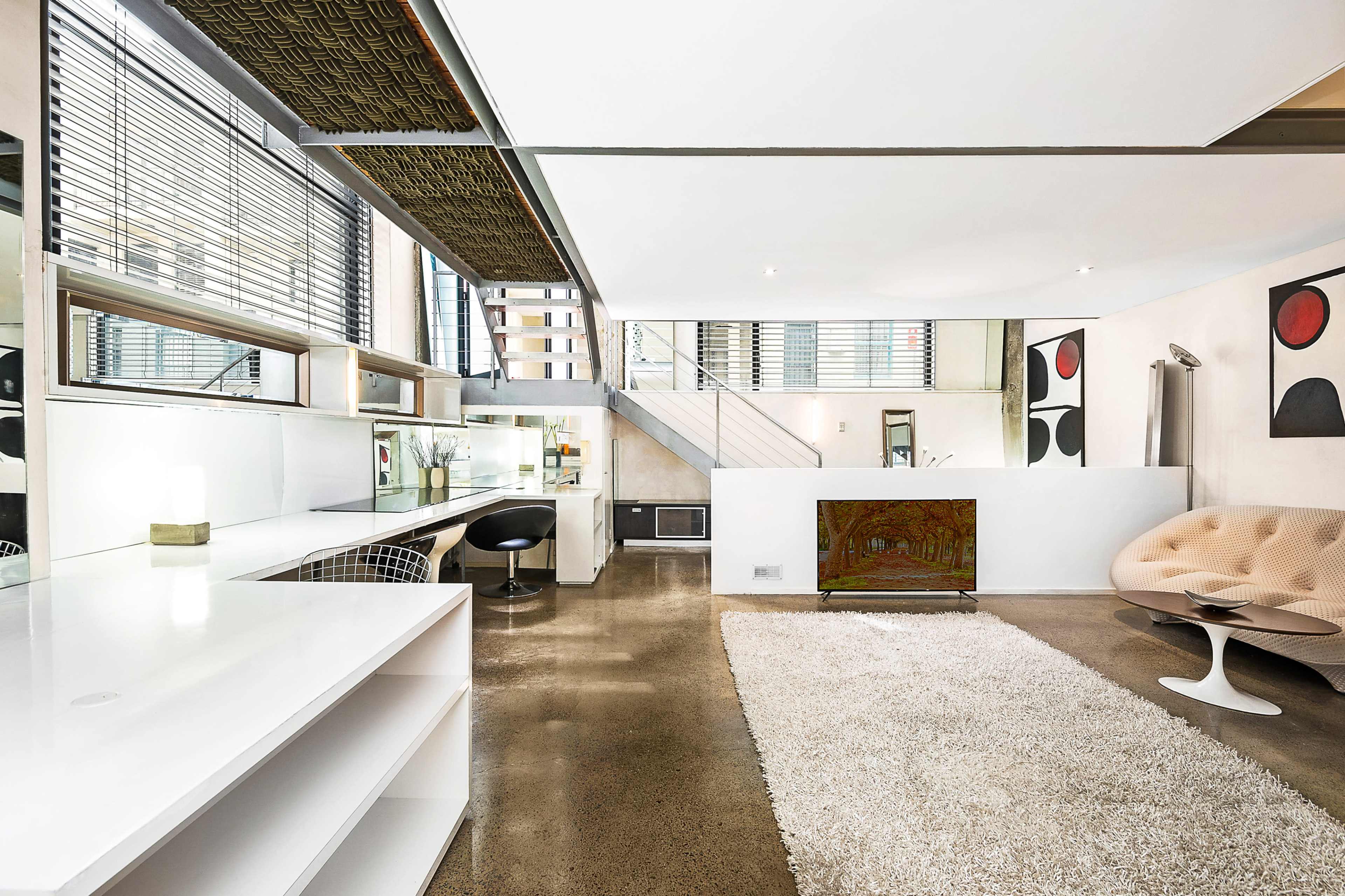 A modern interior space with a glossy white desk, a round beige chair, a partially visible staircase, and large windows allowing natural light to enter.
