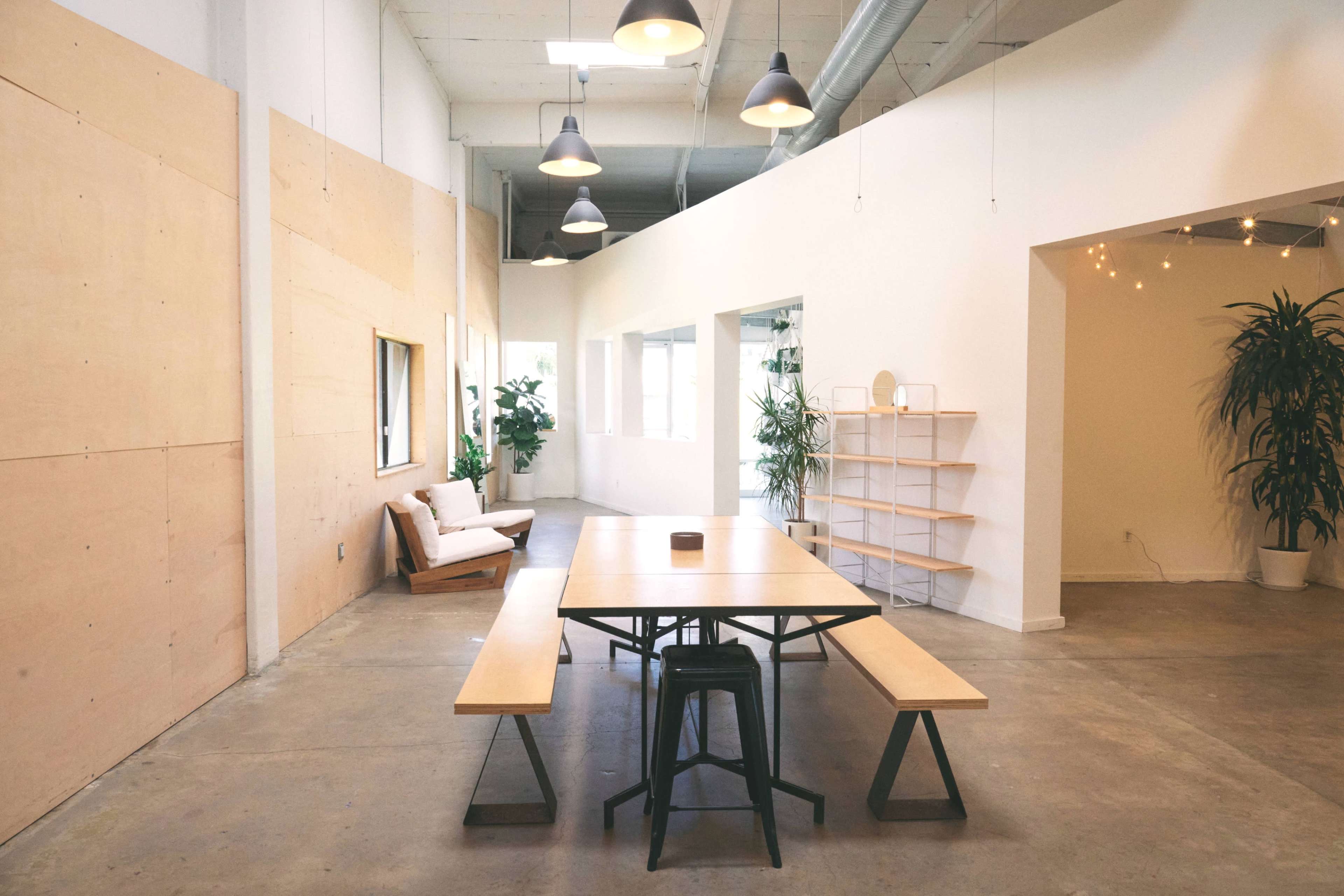 A spacious, well-lit room features a wooden table with benches, surrounded by indoor plants and minimalist decor.