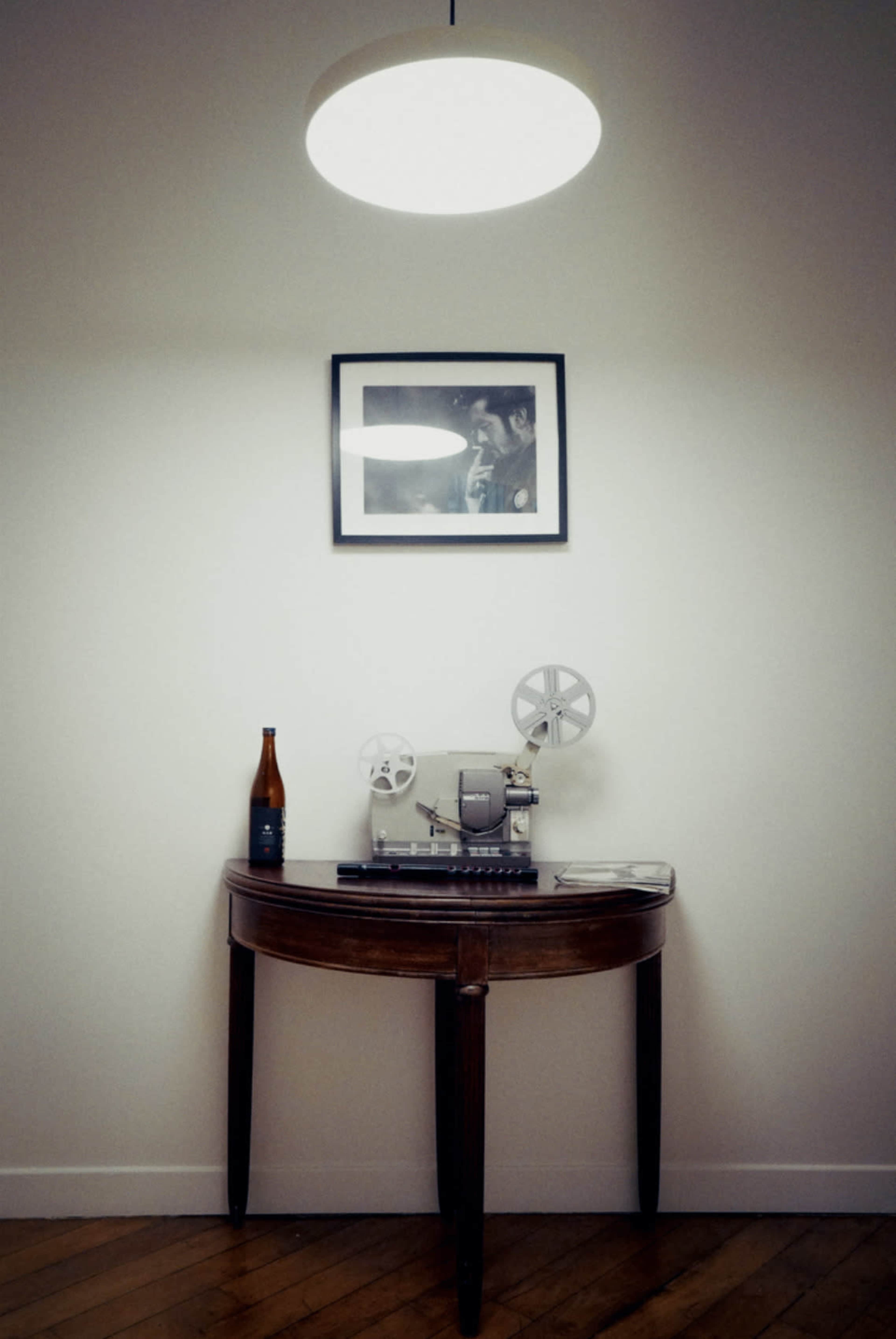A circular light fixture hangs above a round table displaying a film projector, a beer bottle, and a framed photograph on the wall.