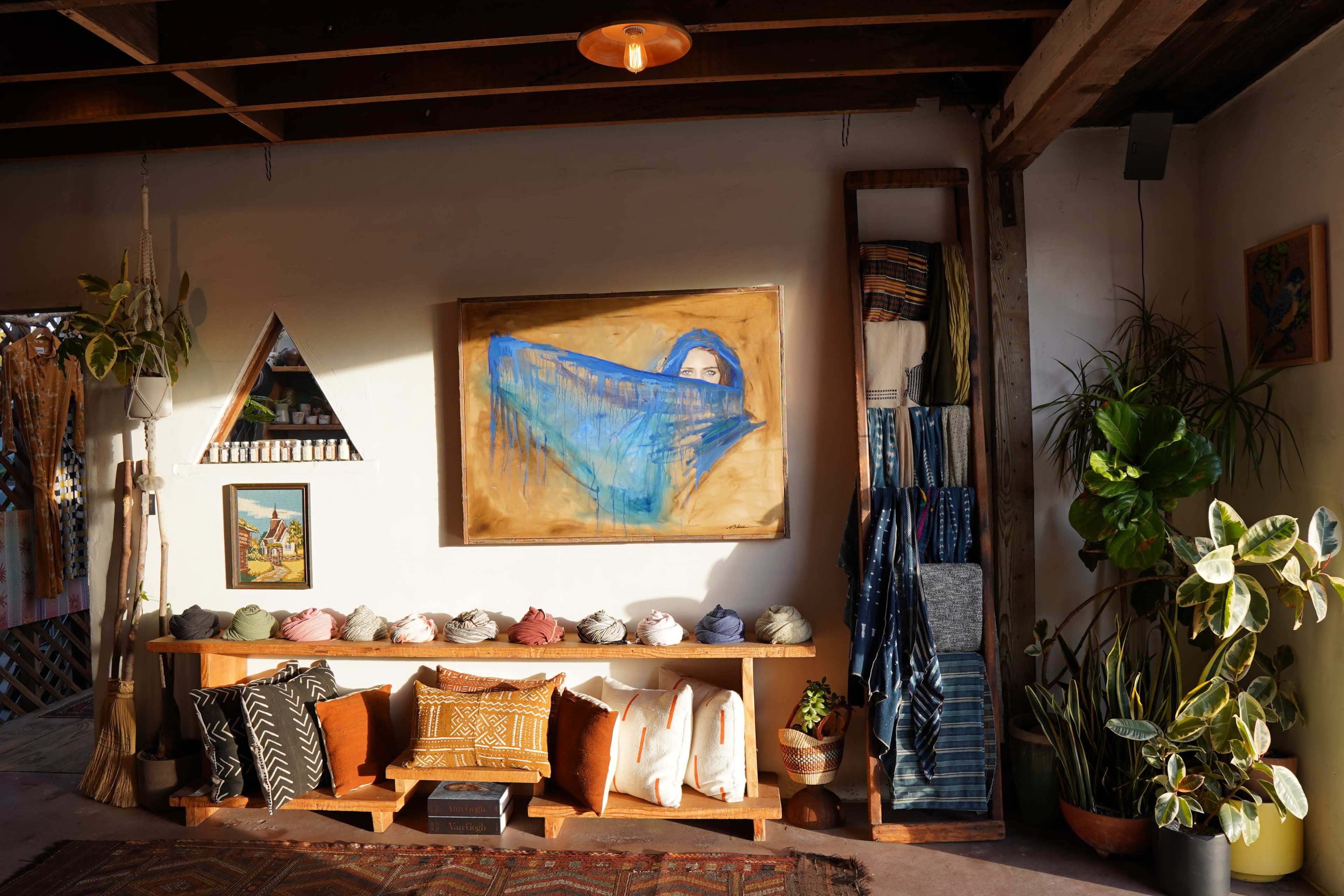 The interior of a shop features a wooden bench with decorative pillows and textiles, a large painting on the wall, and plants arranged around the space.
