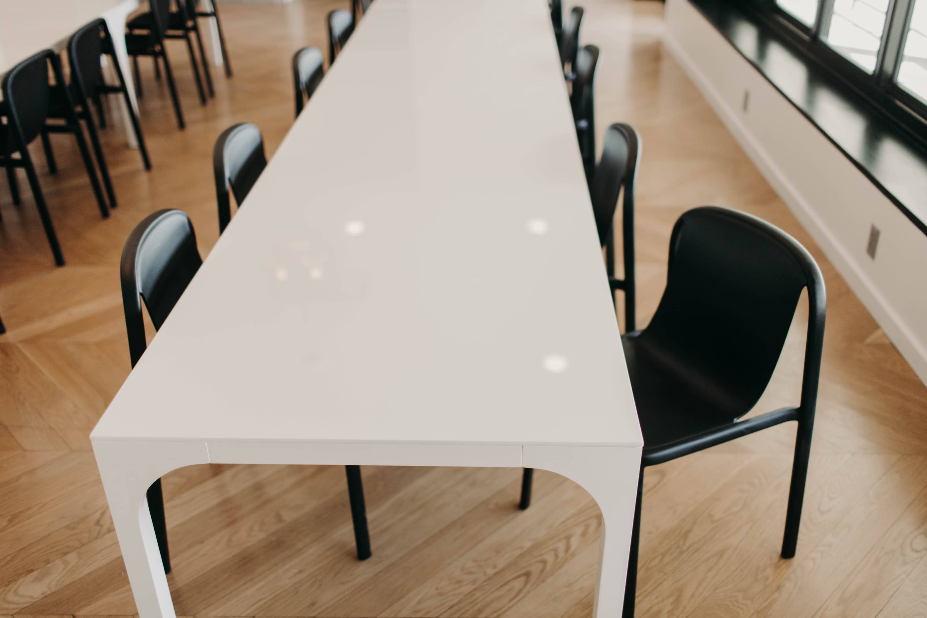 A long white table with black chairs surrounding it is placed on a wooden floor in a well-lit room.