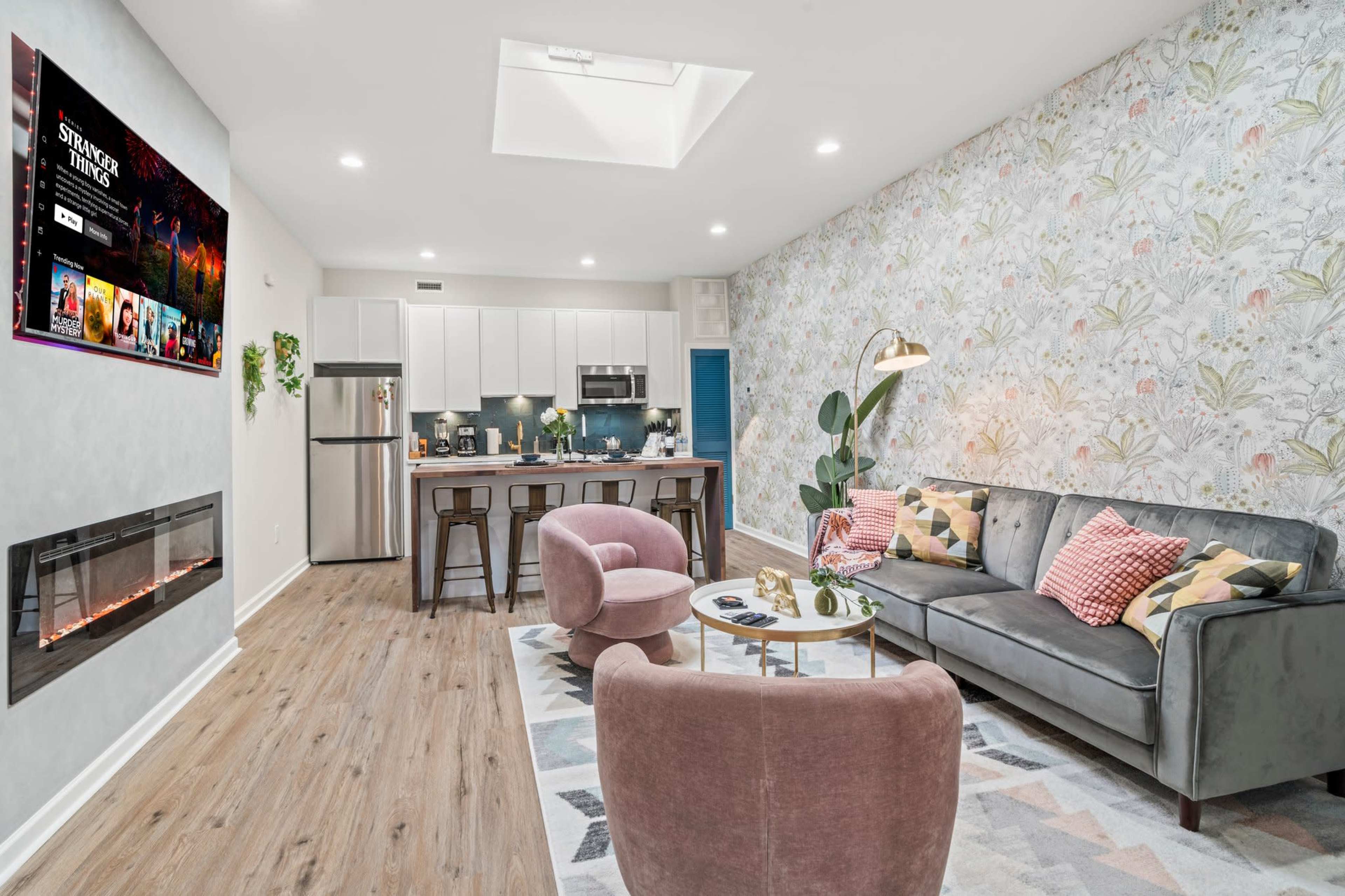 The image shows a modern living room and kitchen area with a television mounted on the wall, a couch, armchairs, a dining table, and a decorative fireplace.