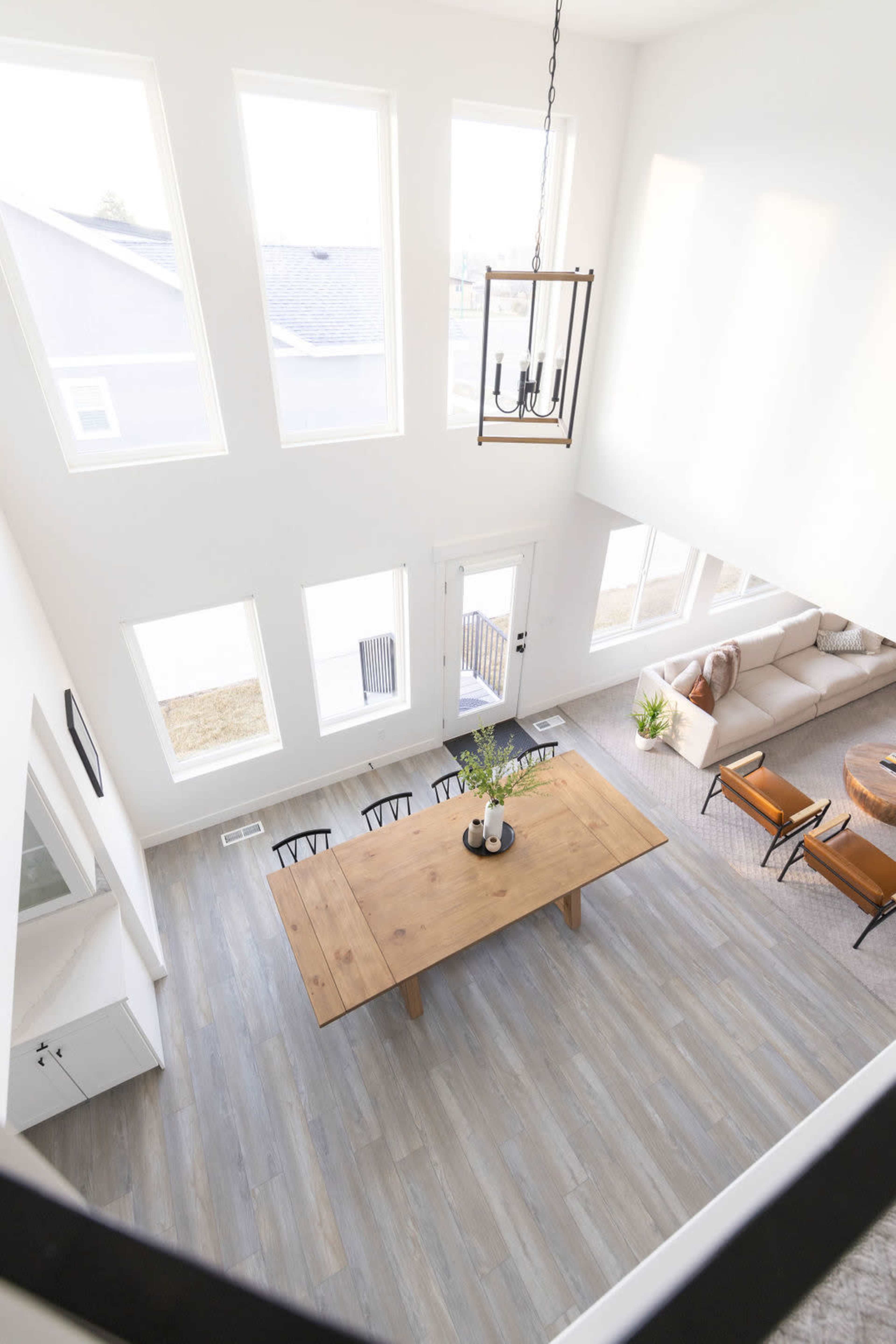 The image shows a large dining table surrounded by chairs in a modern, open-concept living space with high ceilings and tall windows.