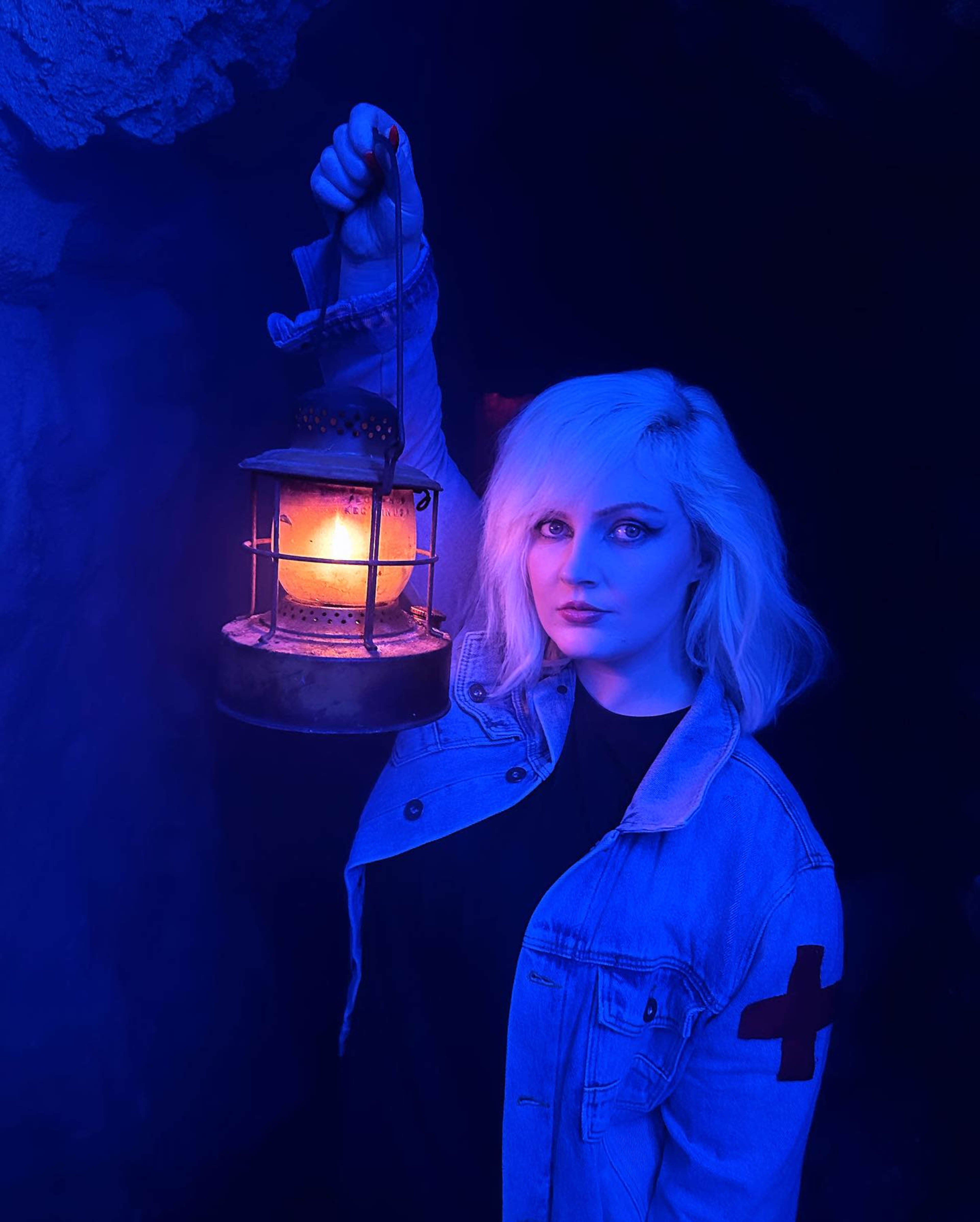 A person with shoulder-length light-colored hair holds a vintage lantern, standing in a dimly lit environment with blue hues.