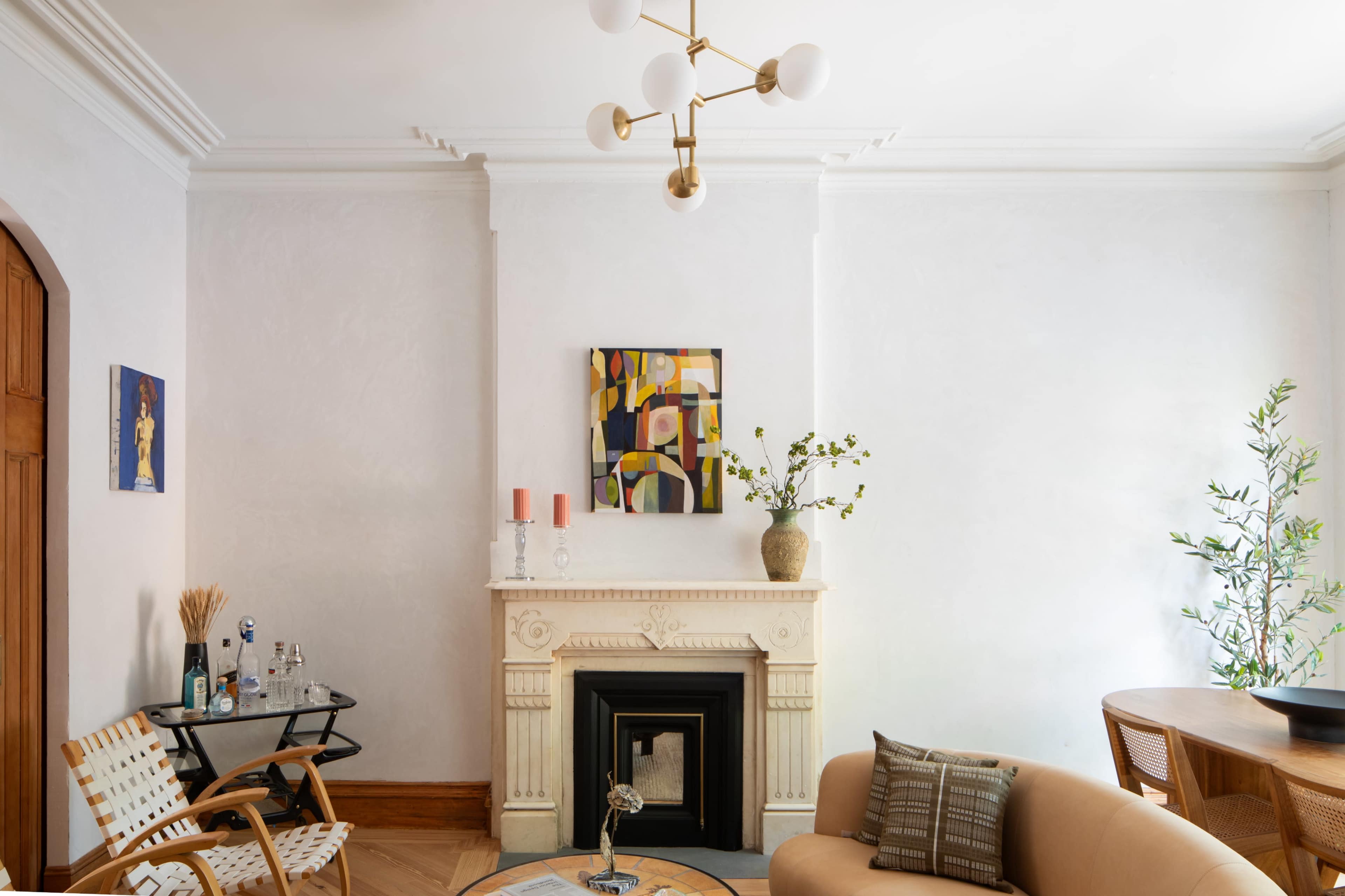The image shows a bright living room featuring a fireplace with decorative molding, a modern light fixture, and a mix of furniture and decor pieces.
