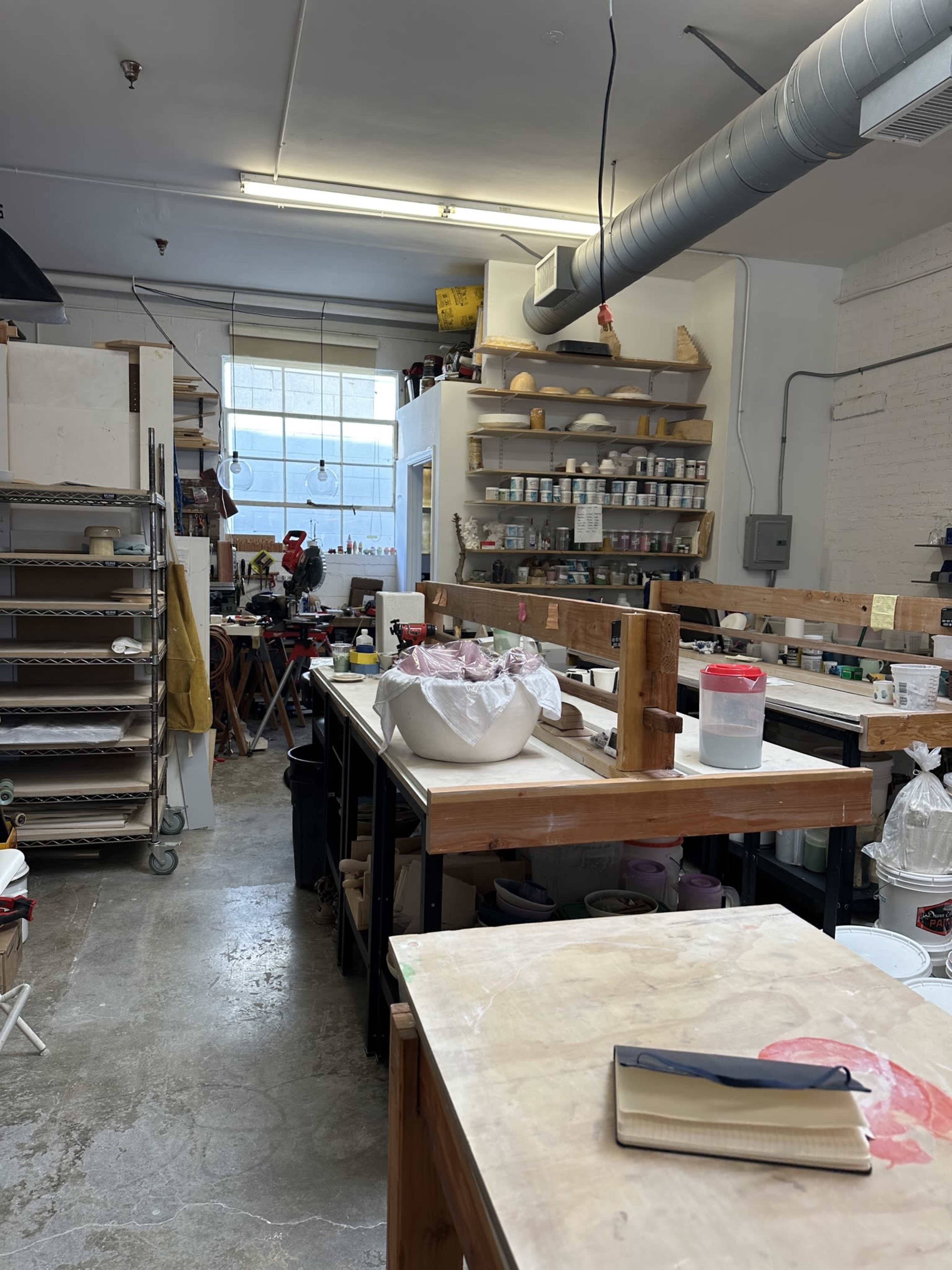 The image shows a pottery studio with various workstations, tools, and materials organized along the walls, all lit by overhead fluorescent lights.