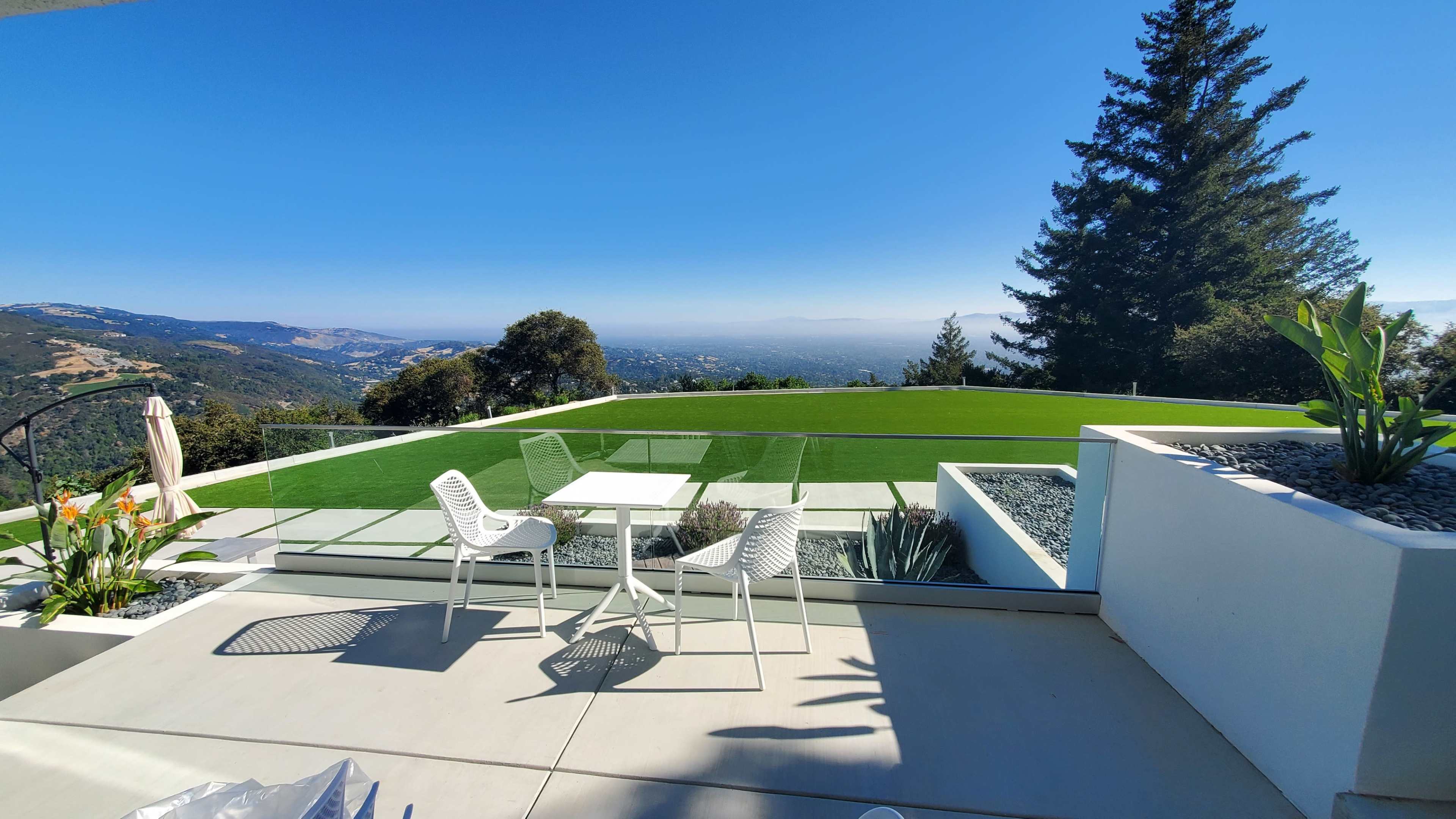 A modern patio features a glass railing overlooking a valley, with a table and chairs set on a concrete surface surrounded by greenery.