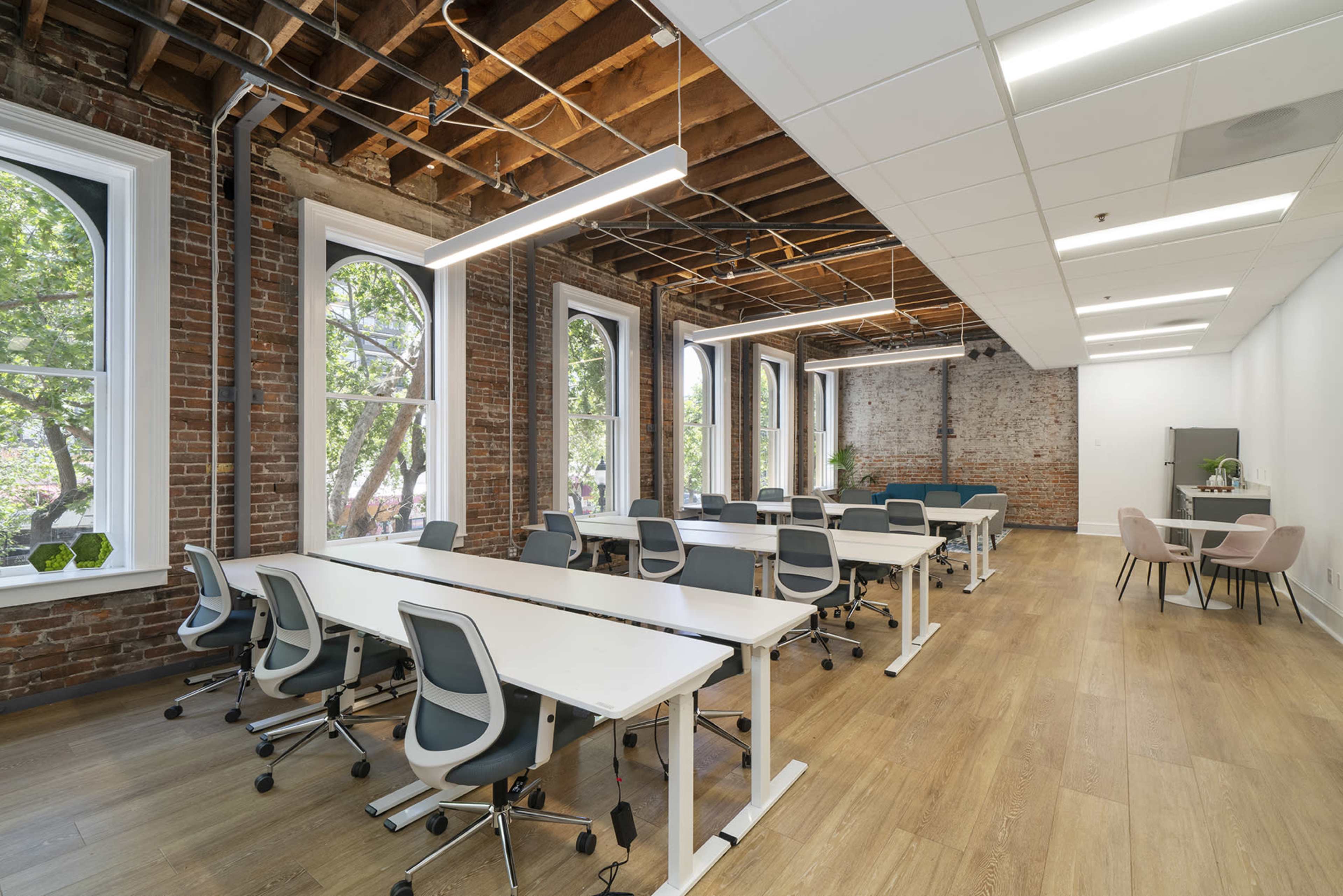 A modern office space features multiple workstations arranged in a bright room with large windows and exposed brick walls.