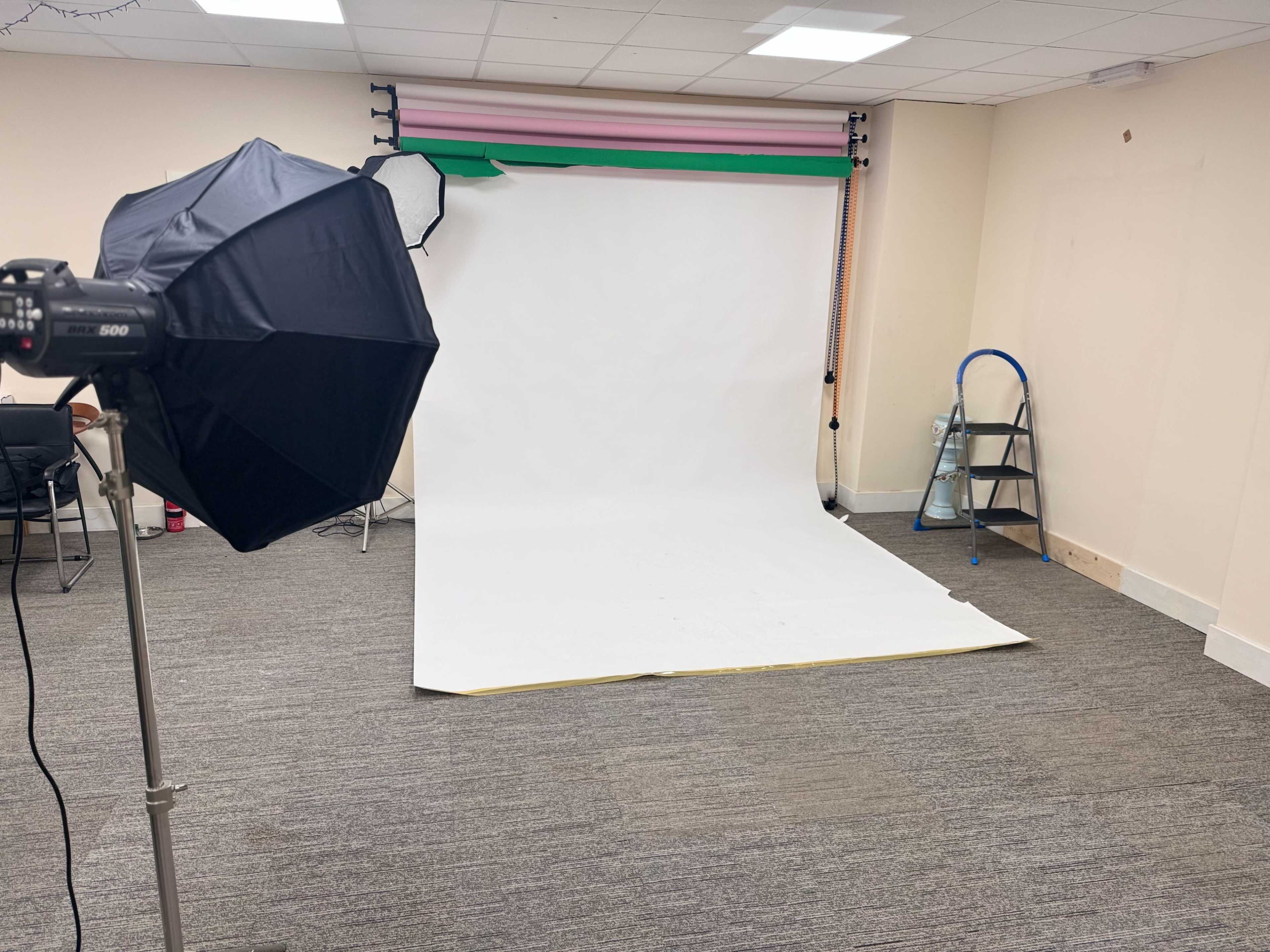 The image shows a photography studio with a white backdrop, lighting equipment, and a ladder in a sparsely decorated room.