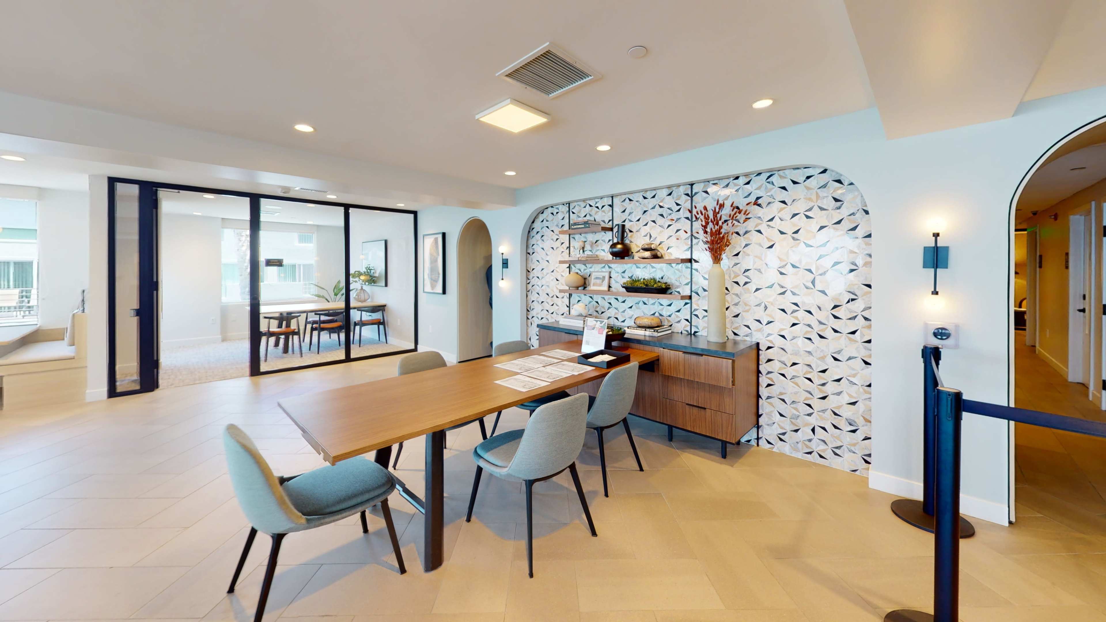 A modern lobby area featuring a central wooden table surrounded by upholstered chairs, with a decorative wall and plants in the background.