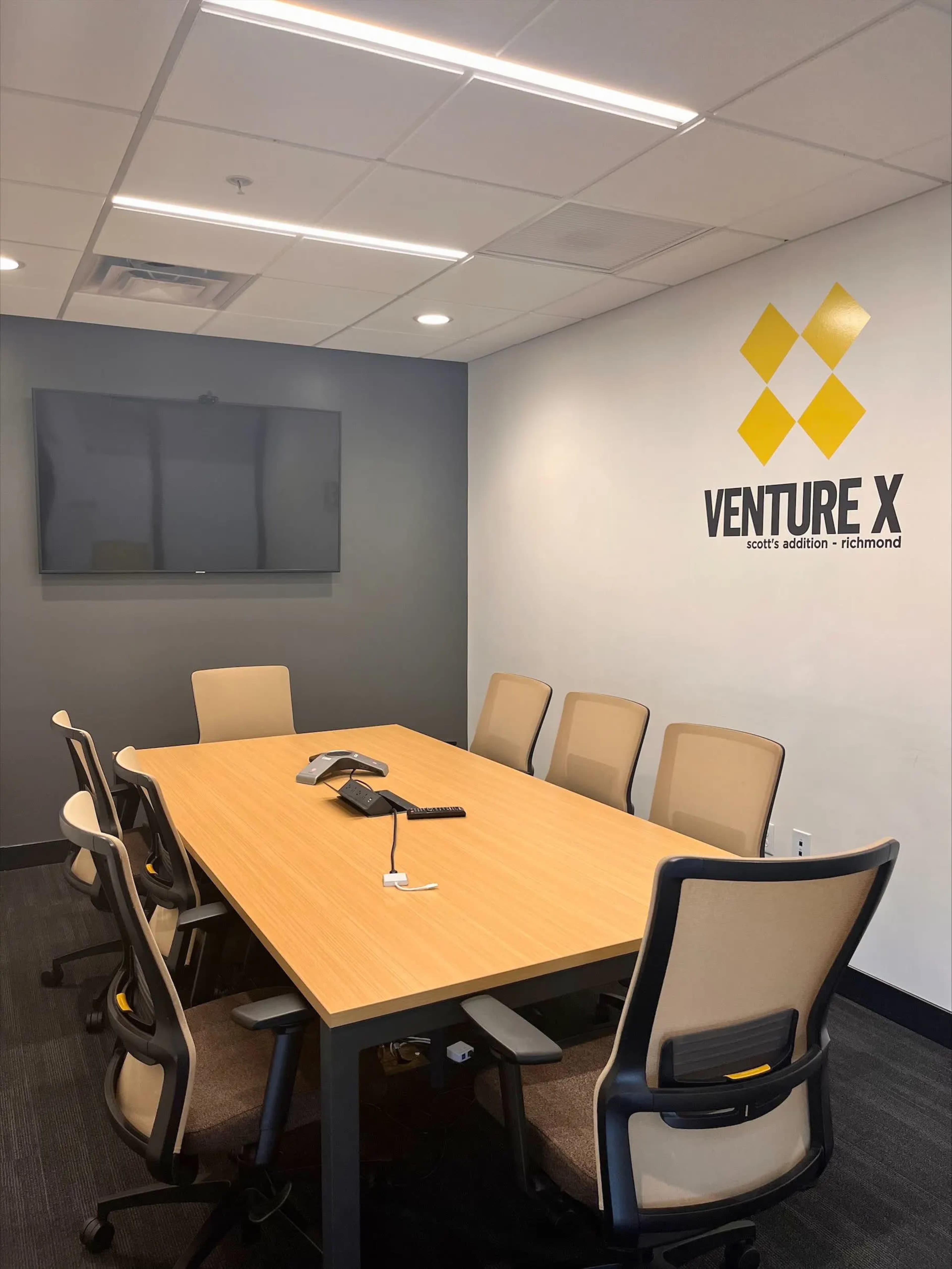 A conference room with a large wooden table surrounded by several chairs, featuring a mounted television and the "VENTURE X" logo on the wall.