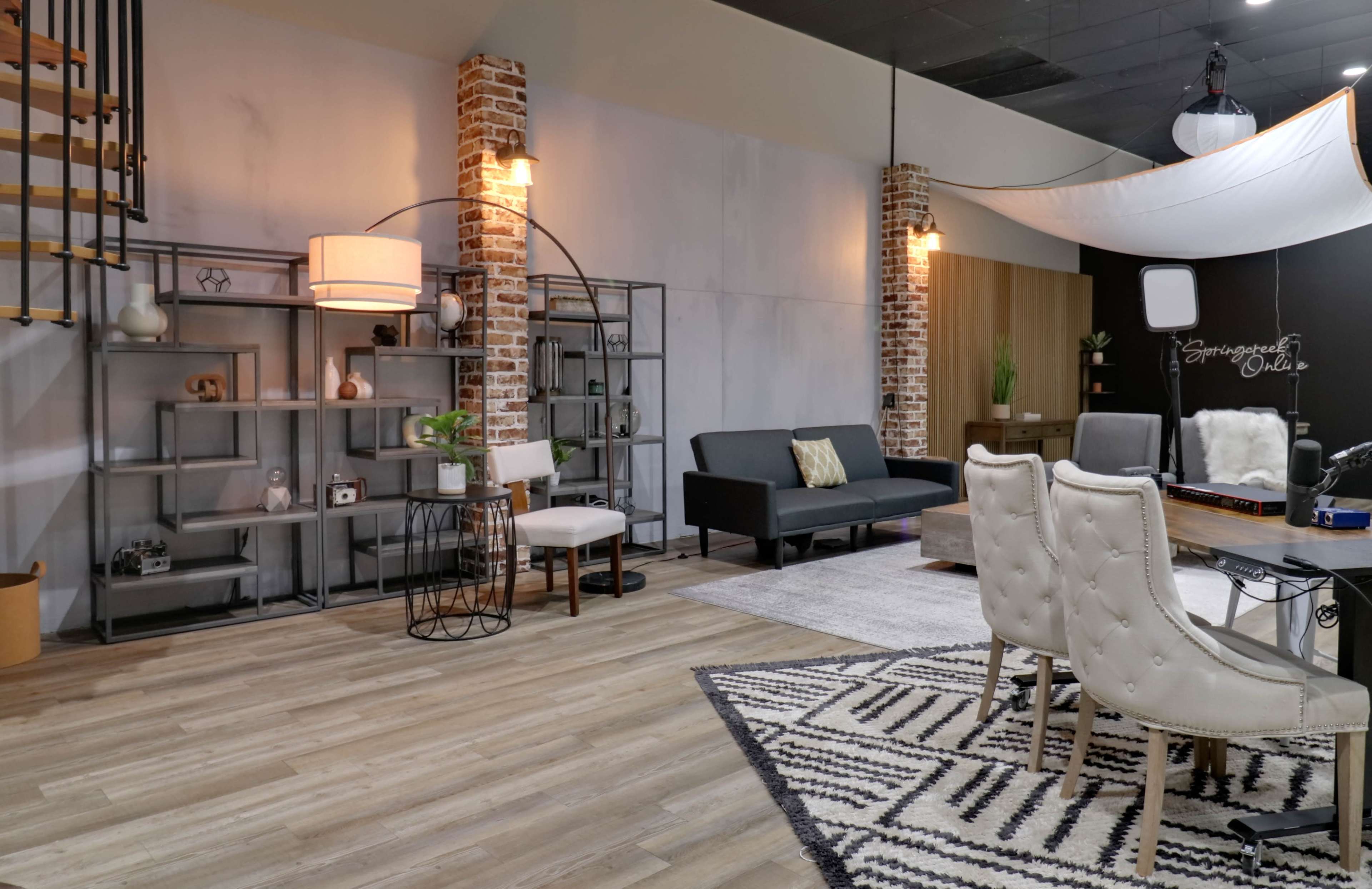 The image shows a modern interior space featuring a mix of industrial and contemporary design elements, with furniture arrangements including a black sofa, dining chairs, and decorative shelves.