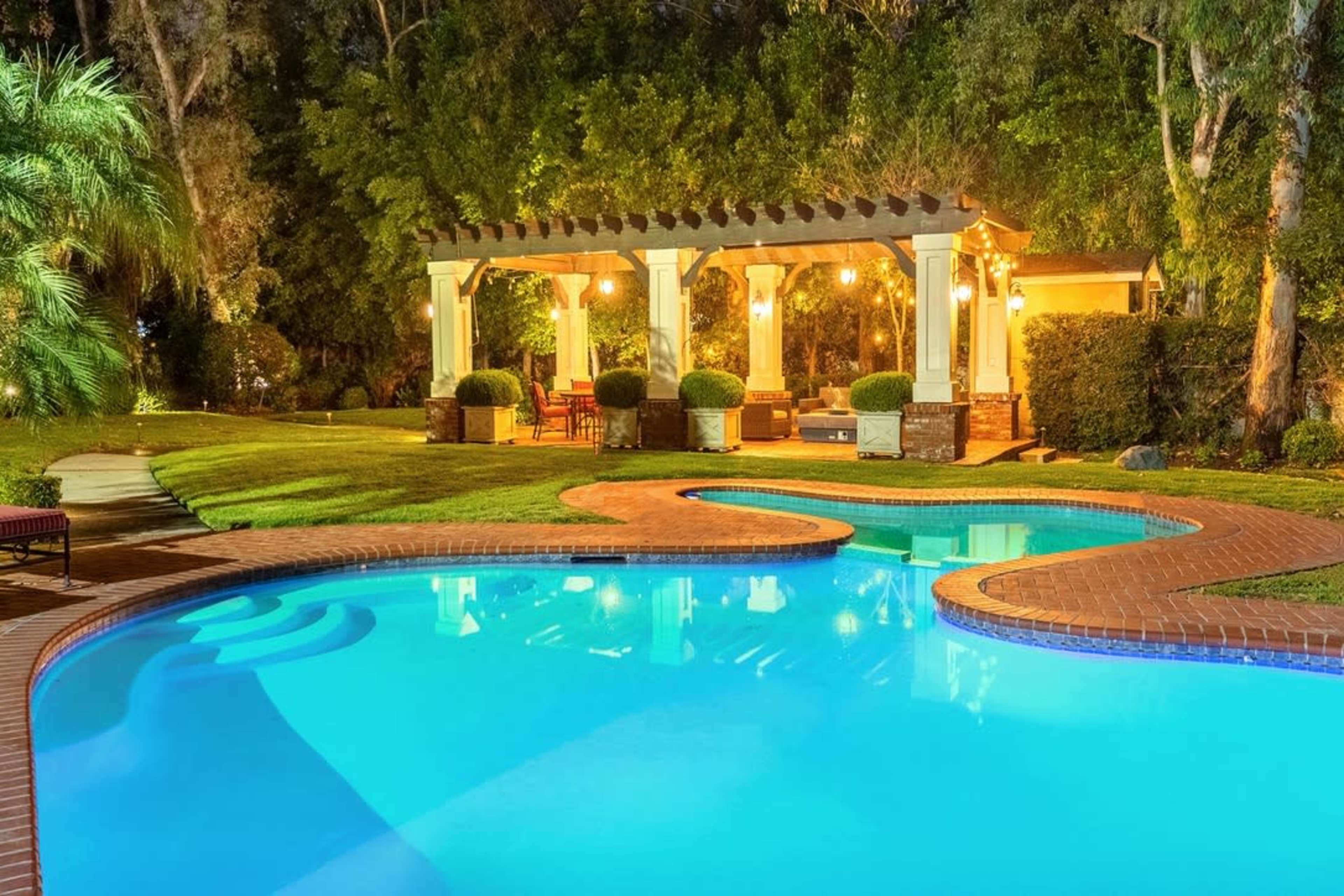 A well-lit outdoor area features a swimming pool with a curved edge, surrounded by landscaped greenery and a pergola with seating.